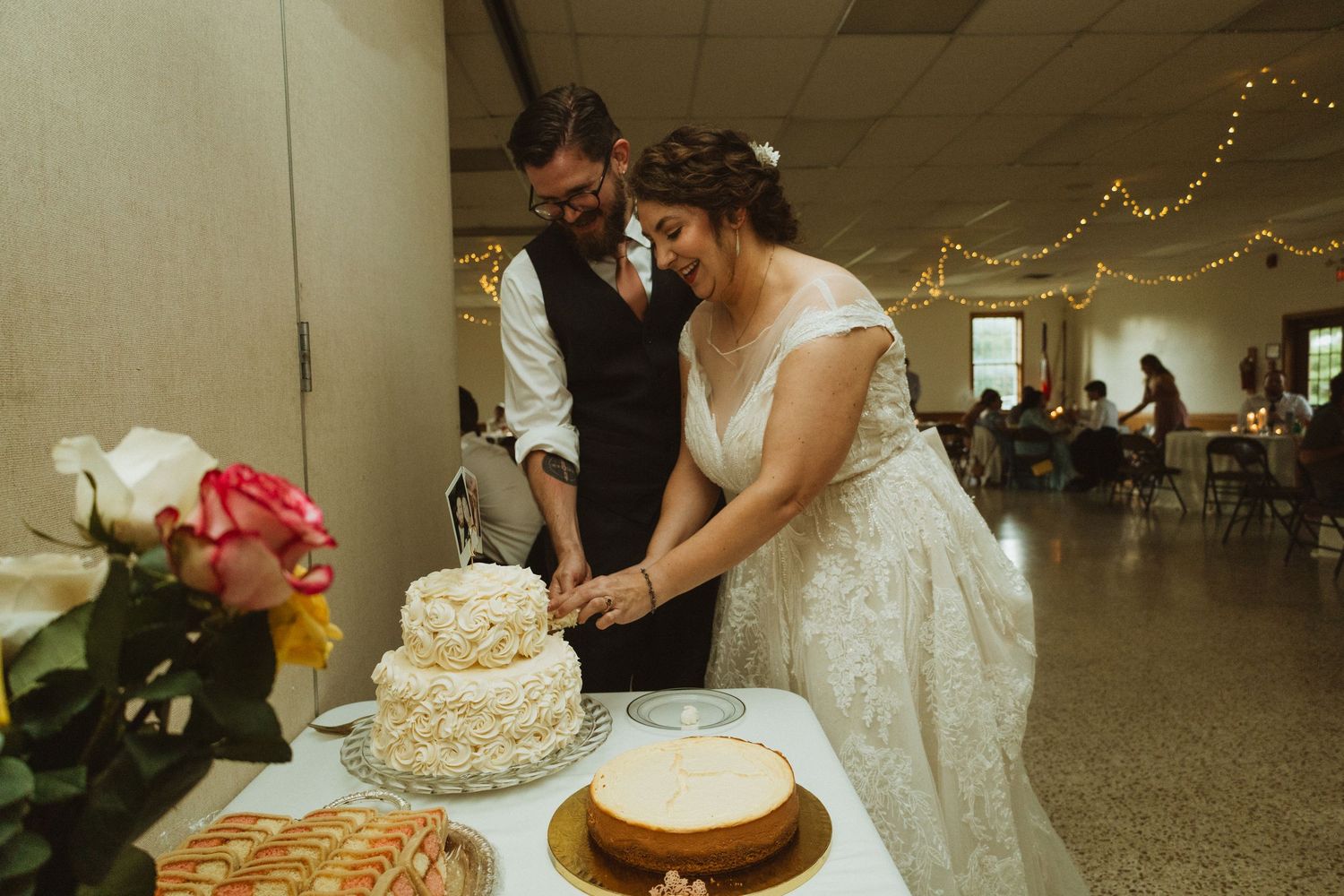 A couple cuts into their multi-tiered wedding cake at a reception with string lights and floral decorations.
