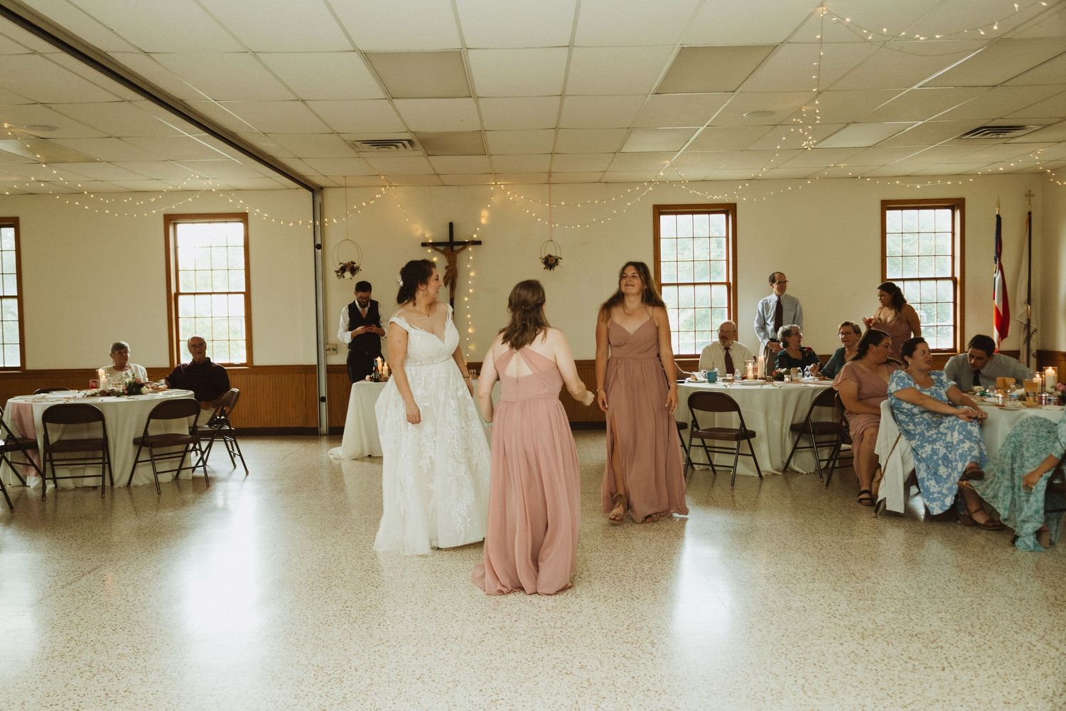Wedding party members in pink bridesmaid dresses gather in a reception hall with folding chairs and tables.
