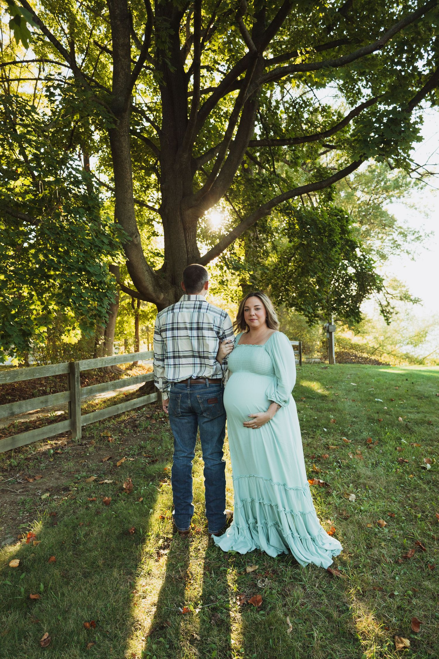 A couple poses together in a sun-drenched outdoor setting with wooden fencing and green trees during a maternity photoshoot.