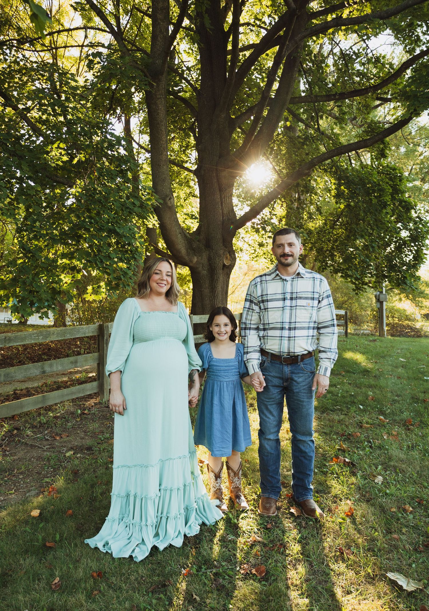 A family stands together in a grassy field at sunset with sunlight filtering through trees in the background.