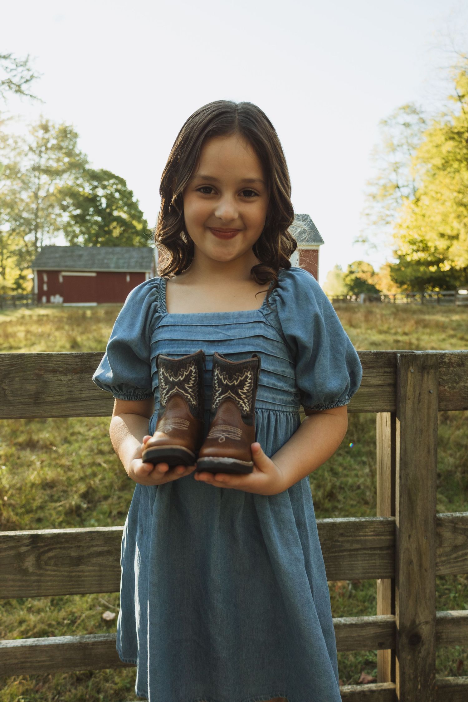 A person in a blue dress stands by a wooden fence holding a pair of small brown leather boots in a rural setting.