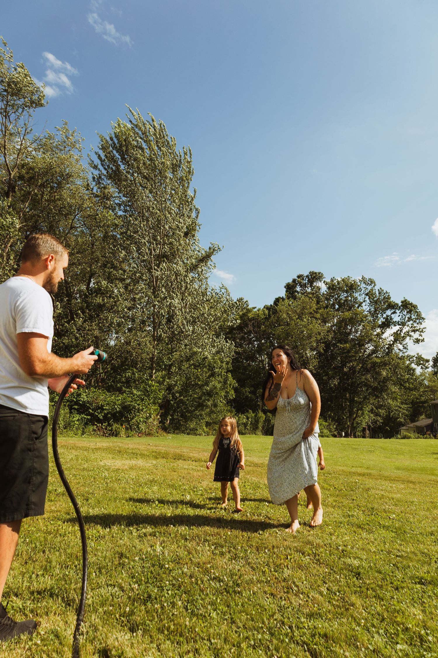 Family enjoying a sunny day outdoors, man holding garden hose while woman and child play on green lawn.