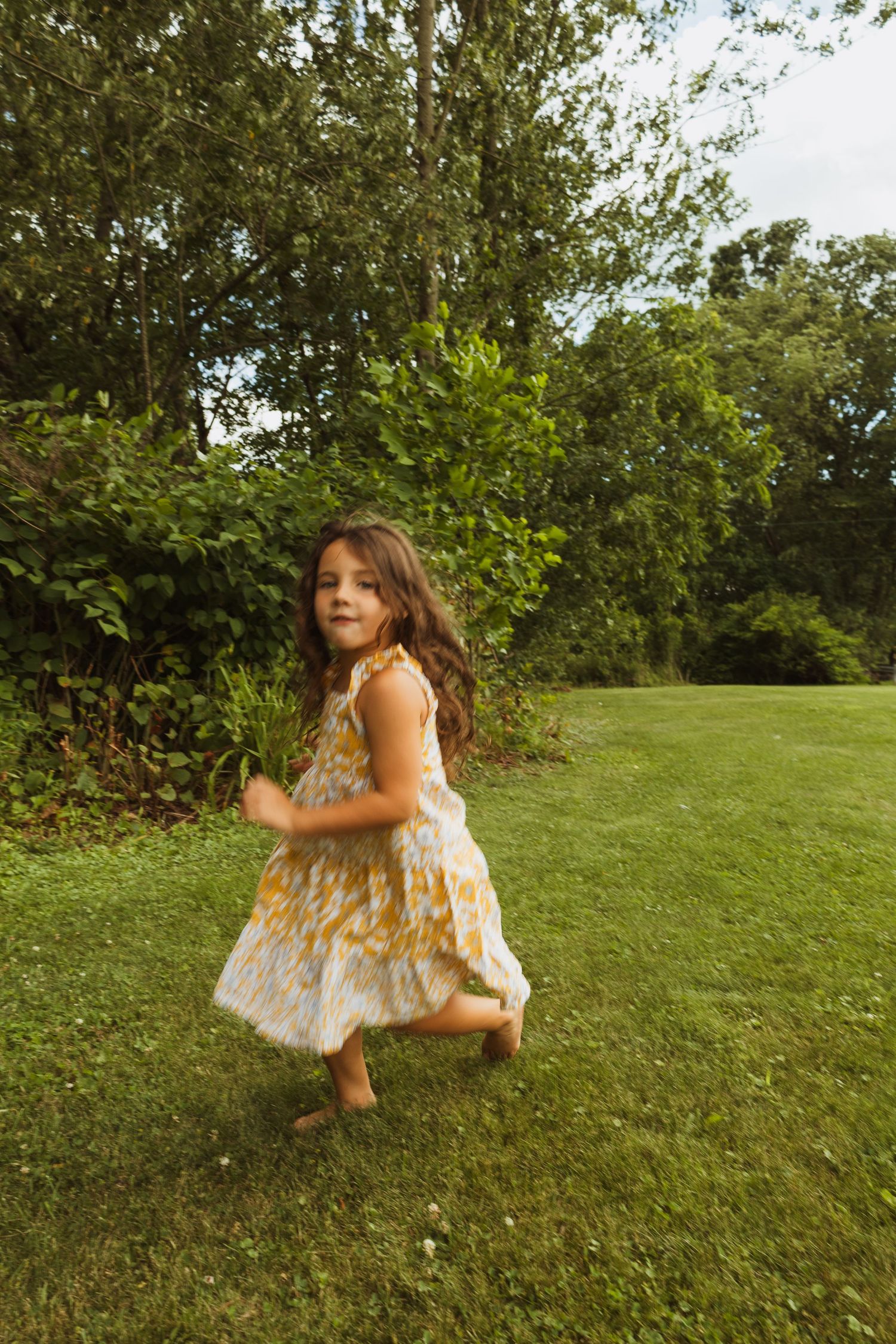 Young girl in floral dress running barefoot on green grass with lush trees in background.