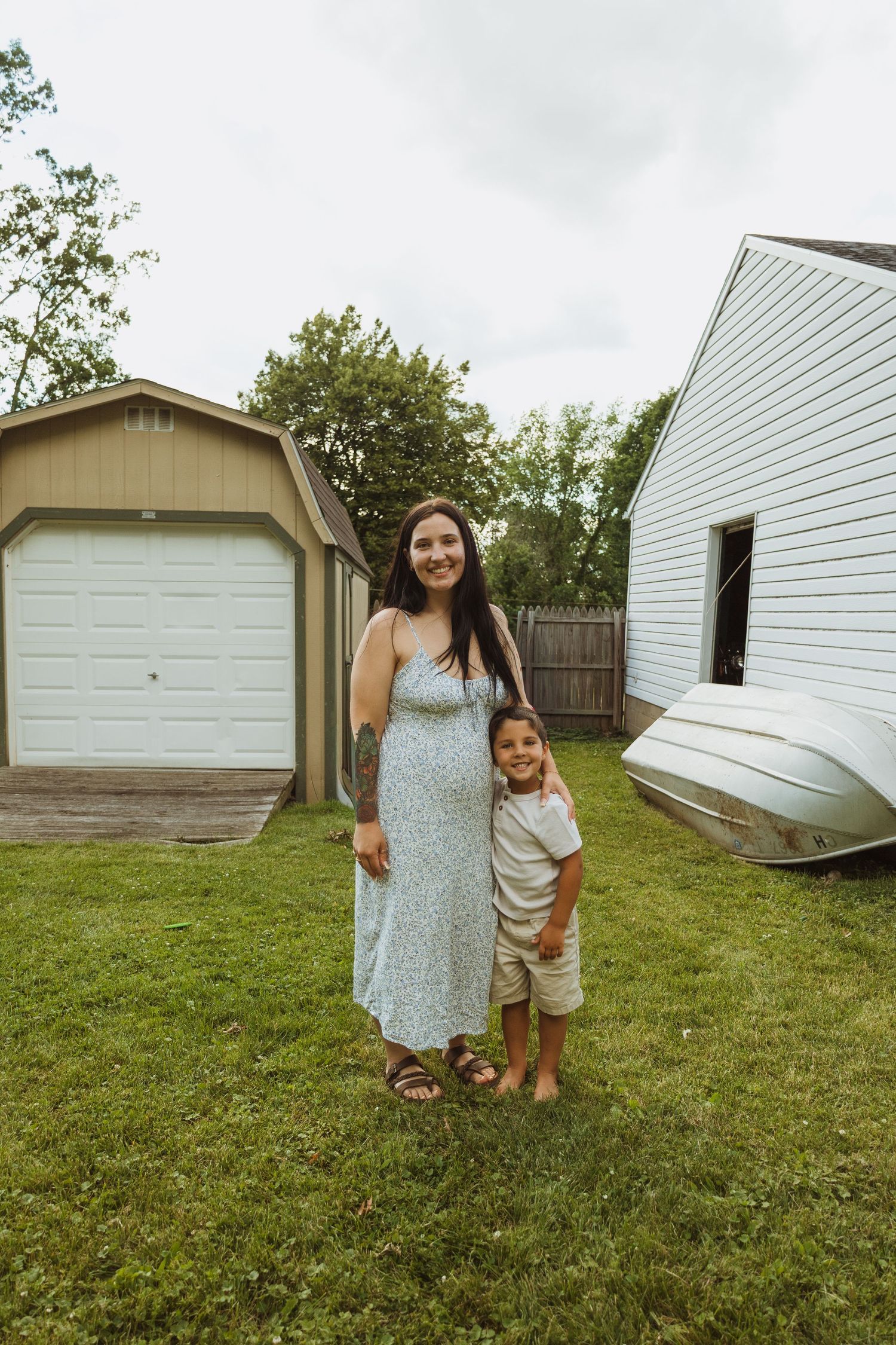 Pregnant woman in blue floral dress poses with young boy in backyard near garage.