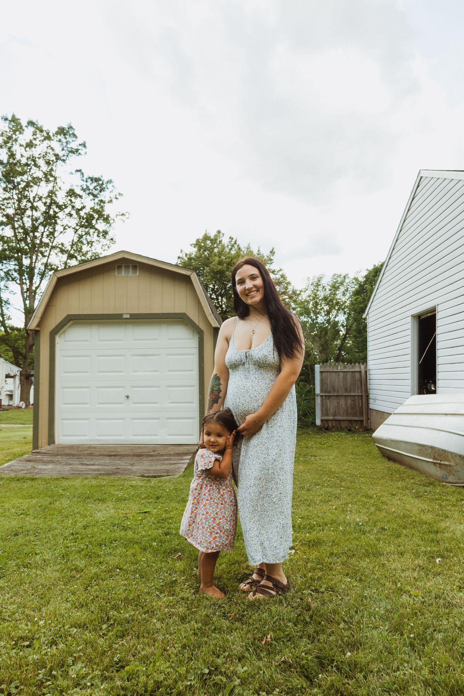 Smiling pregnant woman in floral dress holds hands with young daughter in backyard.