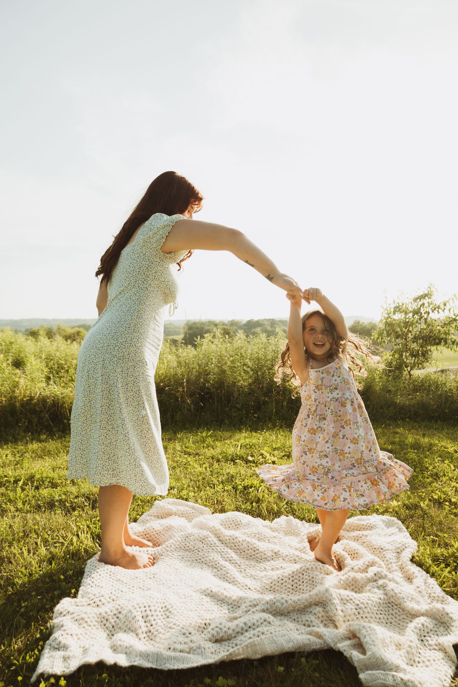 People playing and dancing together on a white blanket in a sunlit meadow.