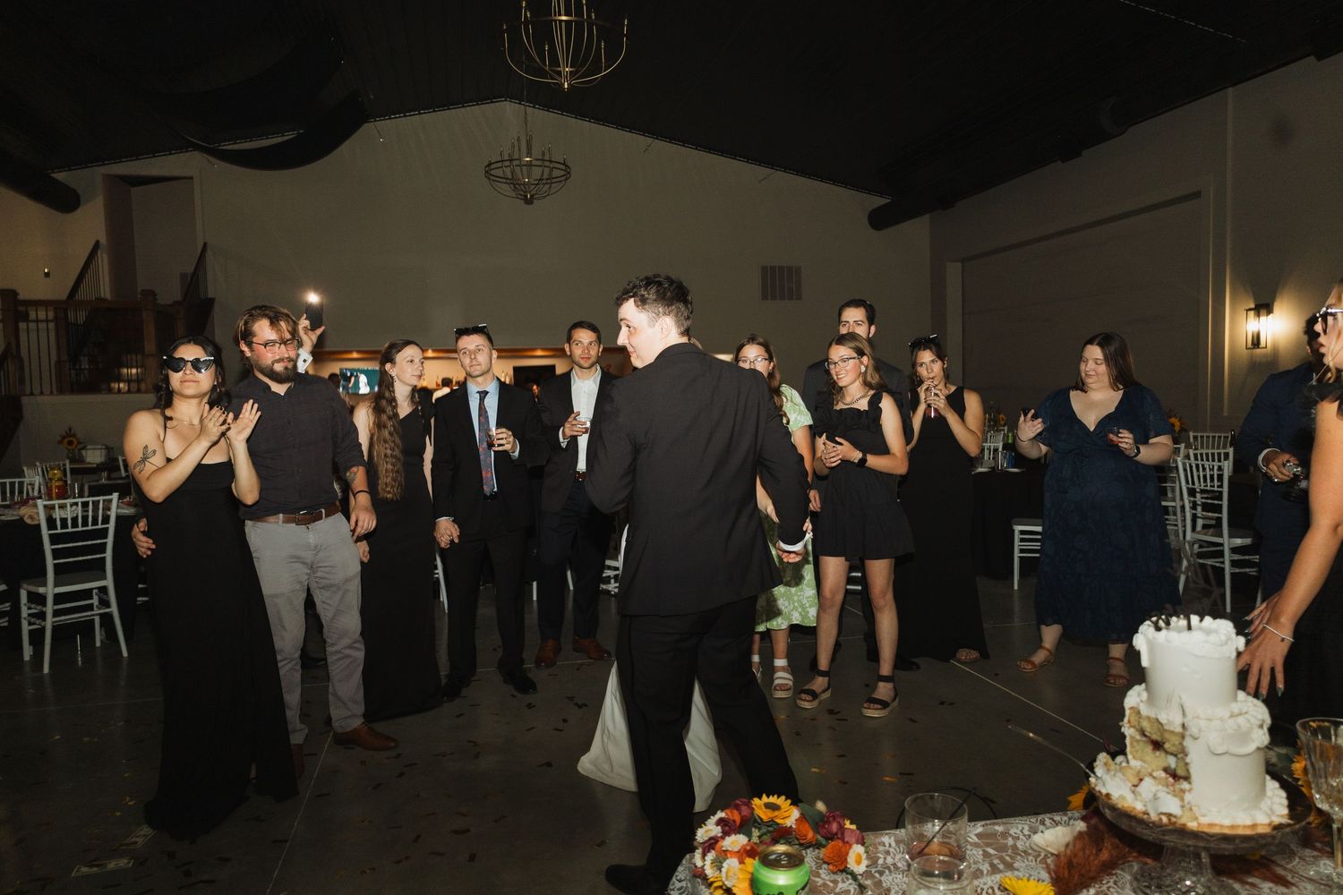 A group of party guests gathered around a wedding cake in a dimly lit reception venue.