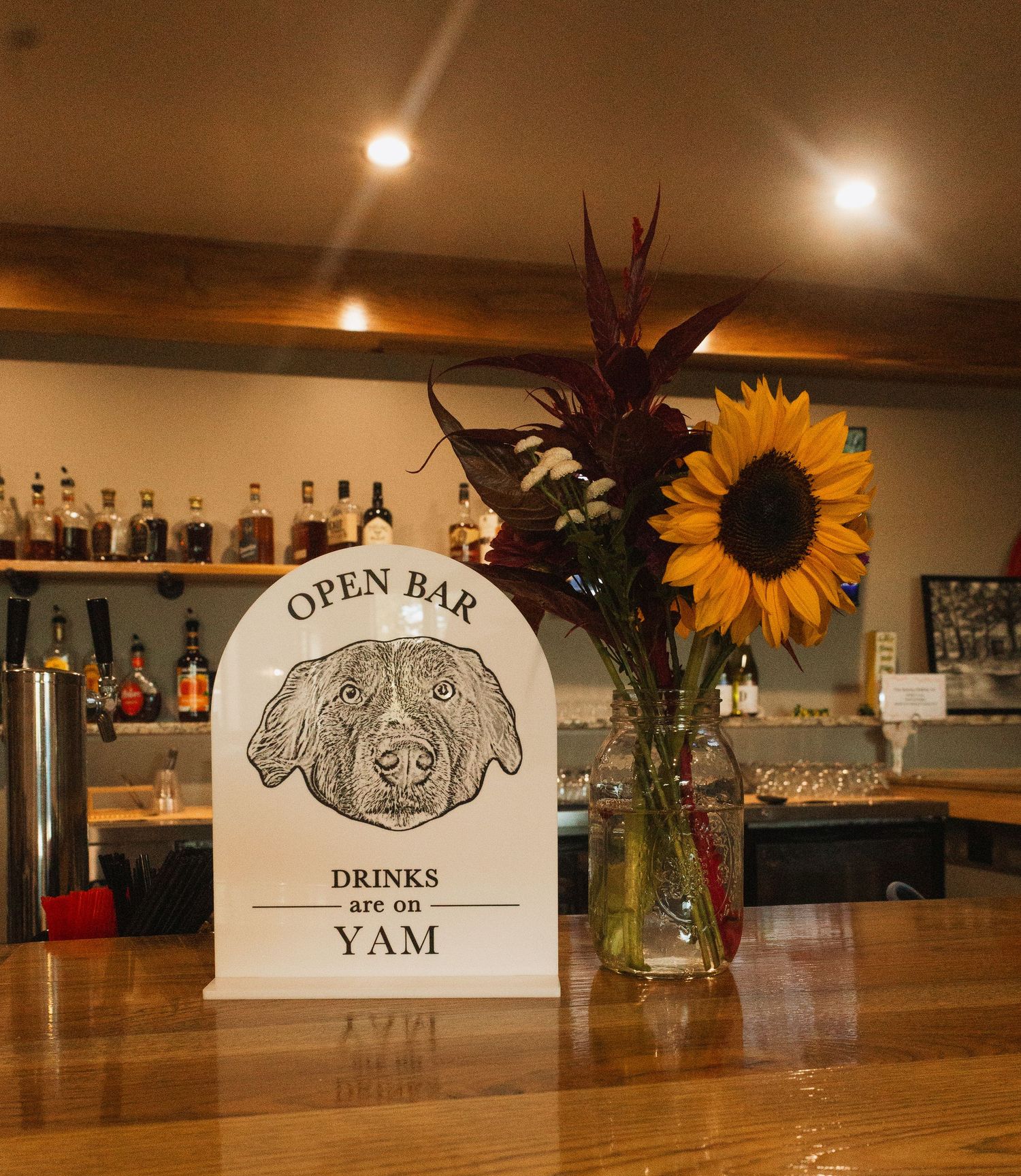 Rustic bar interior with wooden surfaces, sunflowers in vases, and illuminated signage displaying 'Open Bar'.