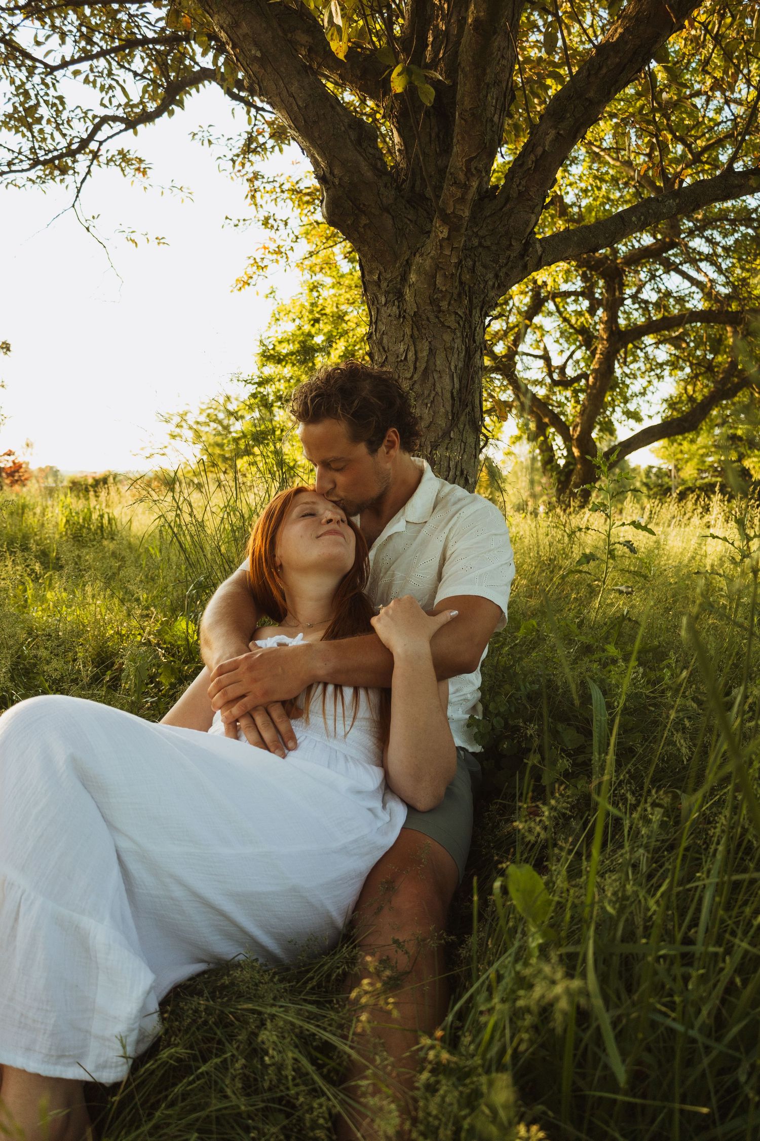 Couple sitting together under a tree in a sunny green meadow, woman in white dress leaning back into mans arms.