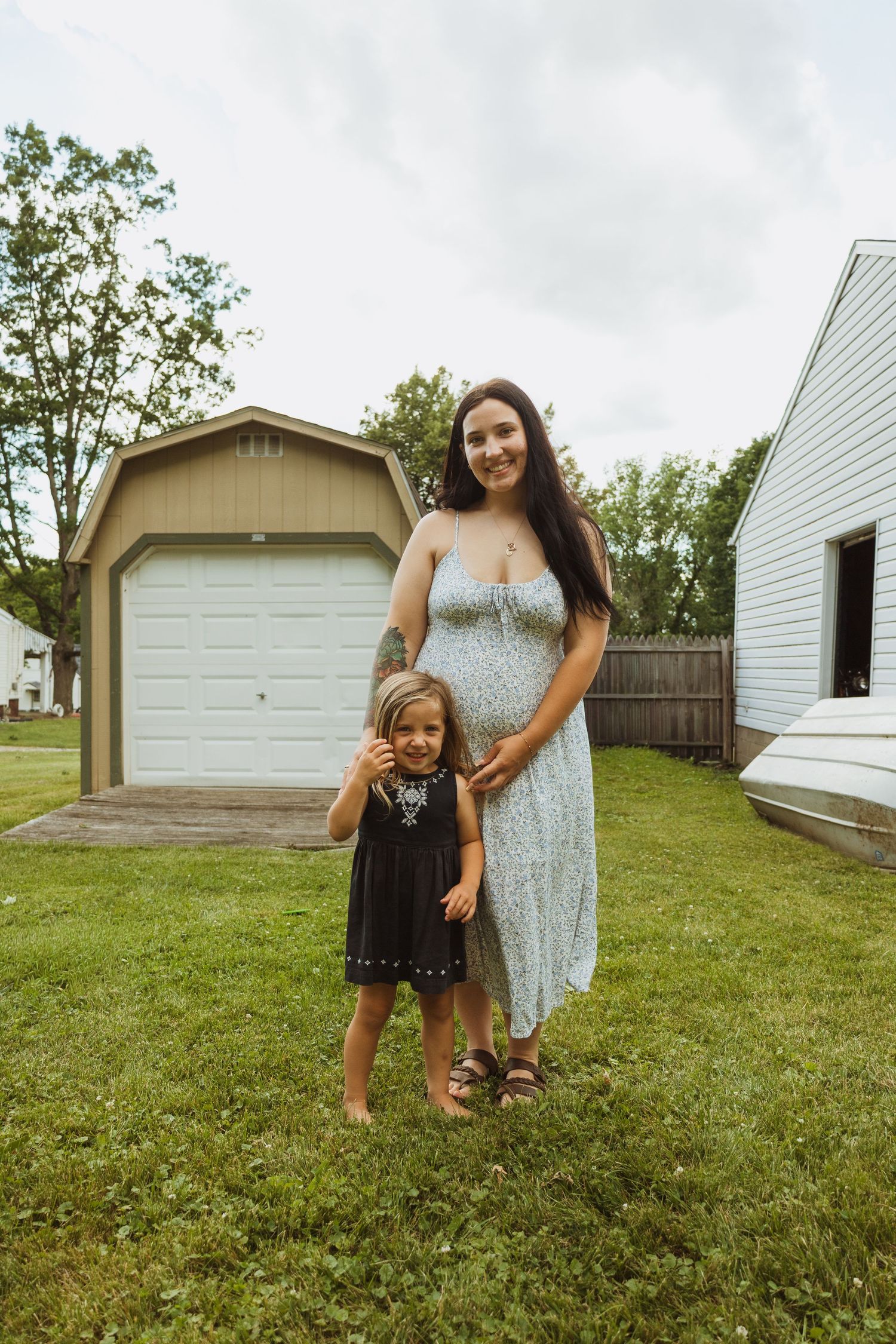 Pregnant woman in floral dress stands with young child in black dress on green lawn near garage.
