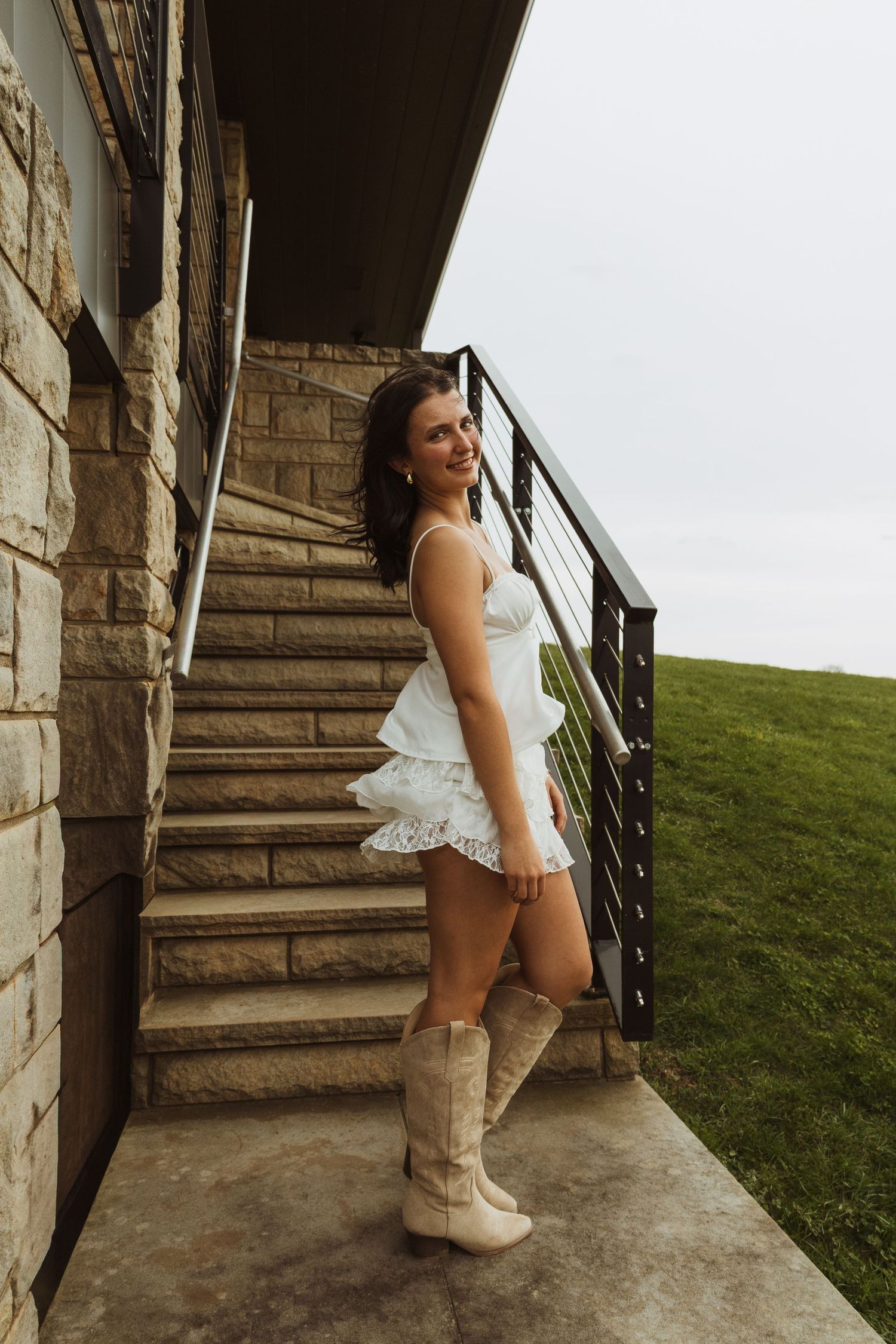 Young woman in white ruffled mini dress and cowboy boots posing on stone steps outside a building.
