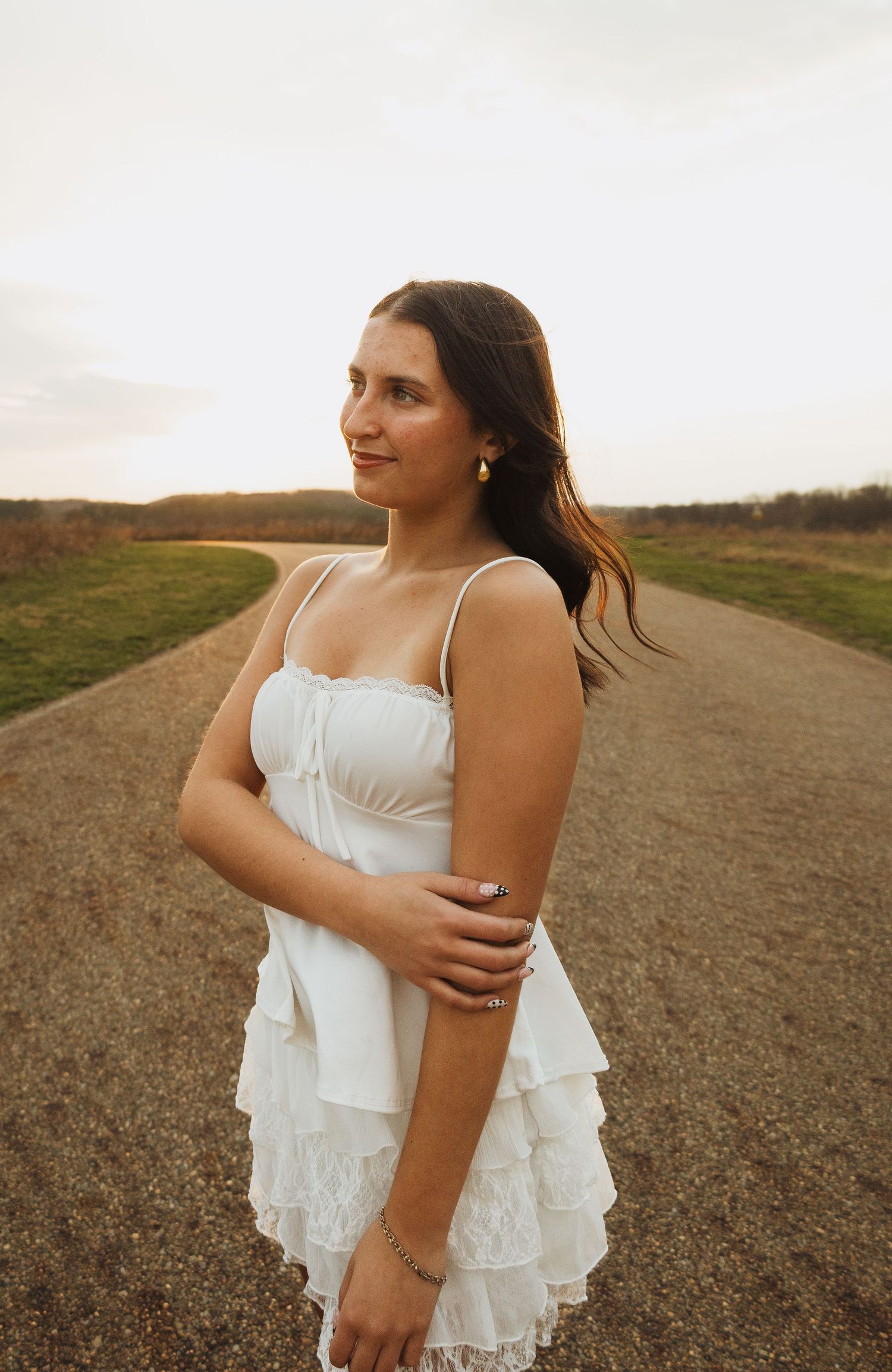 Young woman in white ruffled sundress standing on a country road at sunset, looking gracefully to the side.