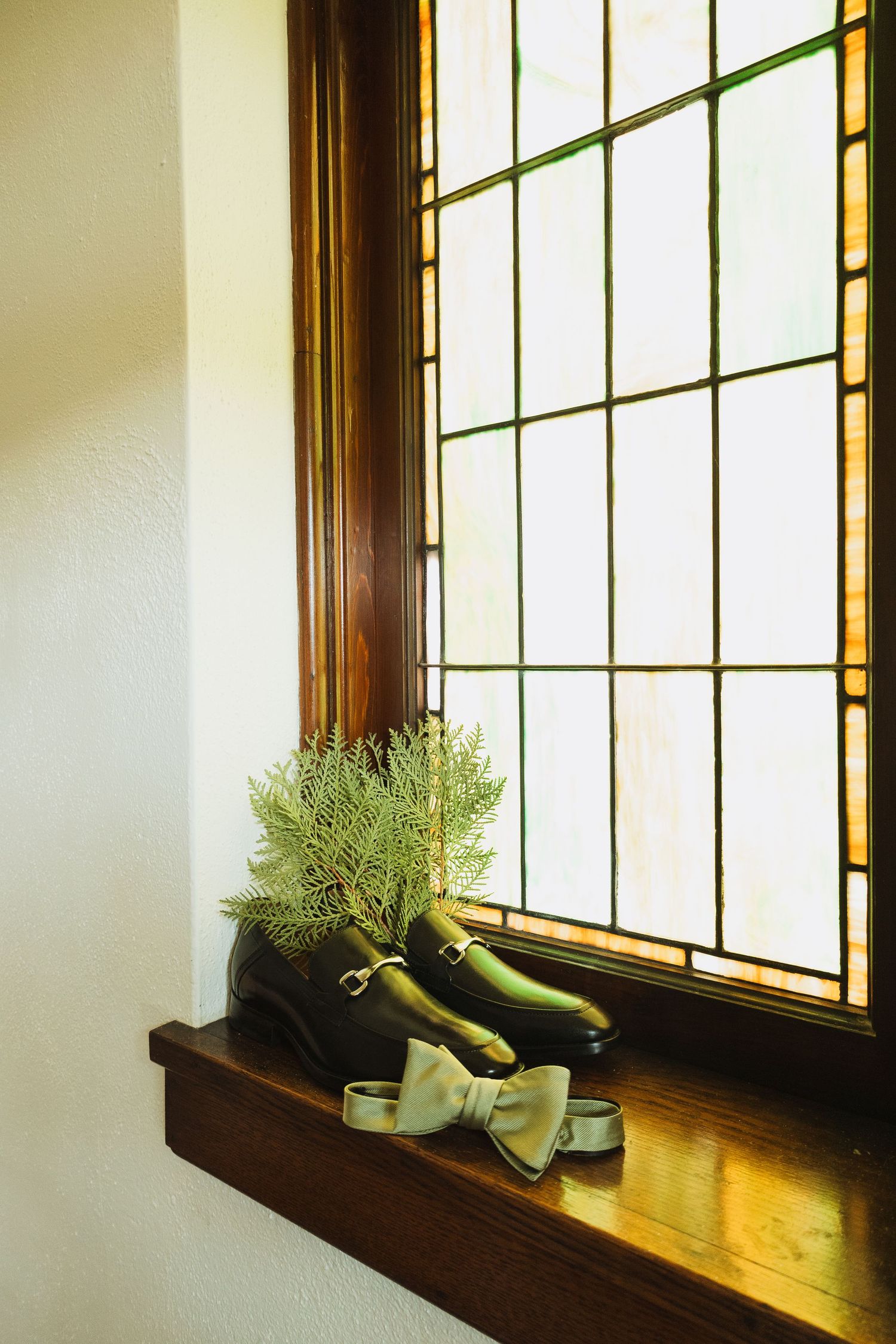 Black loafers with bow tie and greenery displayed on wooden windowsill beside stained glass window.