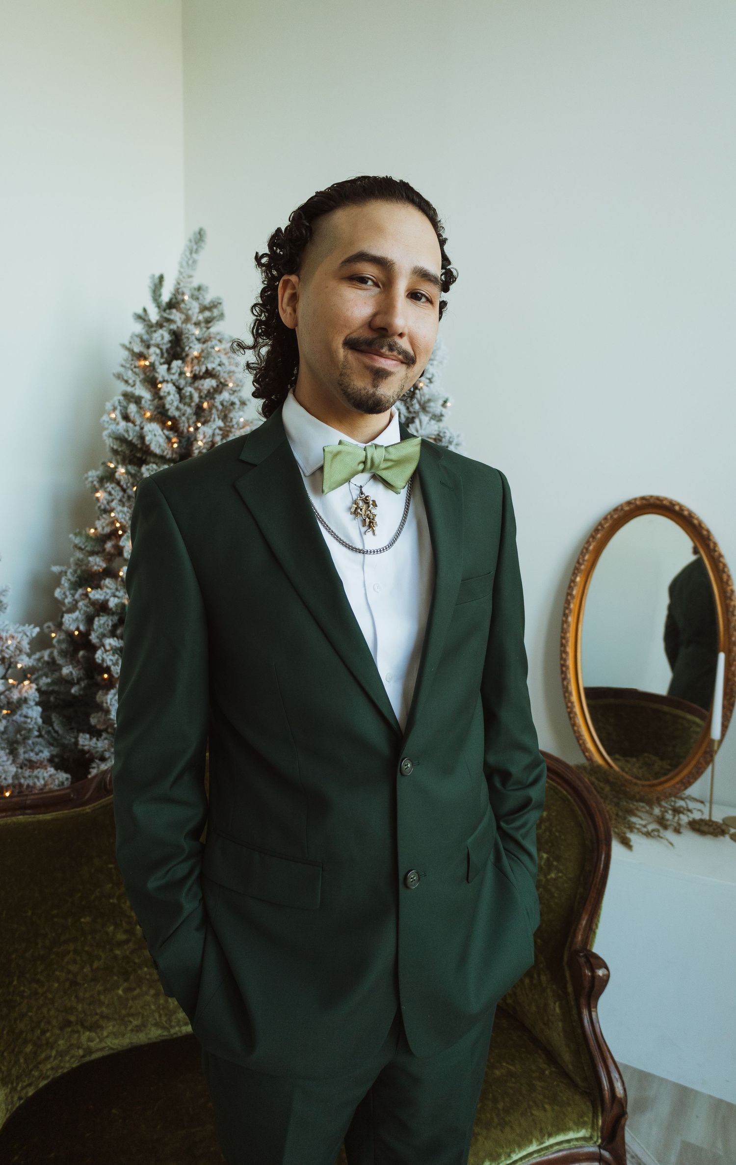 Groom in dark green suit with sage bow tie smiling before Christmas tree in elegant room.