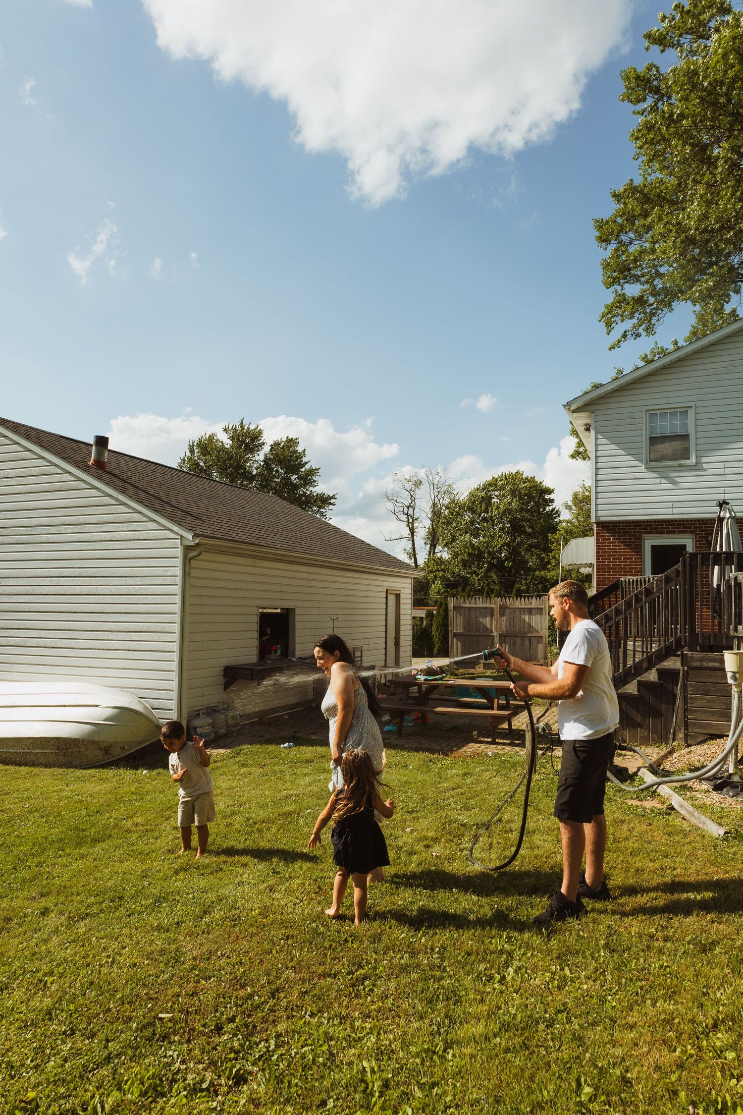 Family enjoying backyard fun with a garden hose on a sunny day near a white house with green lawn.