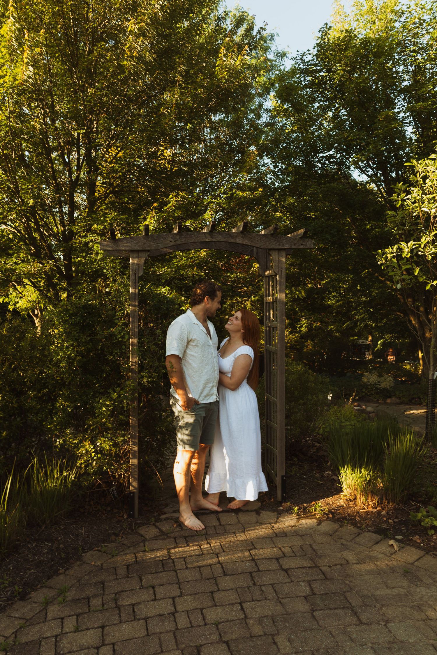 Couple standing under a wooden garden arbor surrounded by lush green trees, sharing a romantic moment on a sunny day.