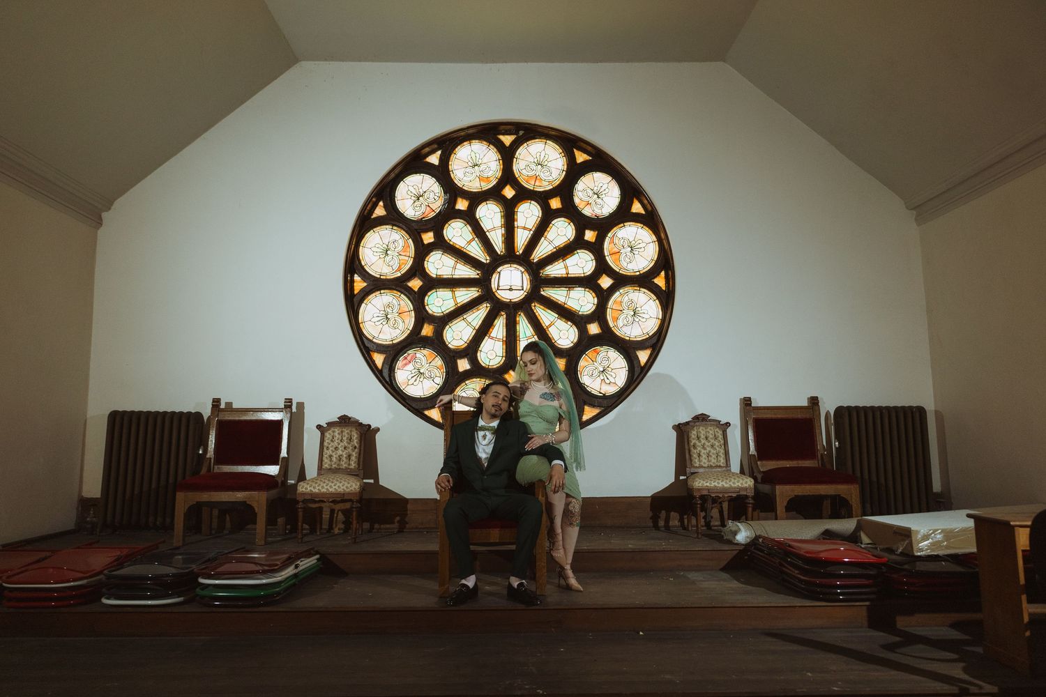 Bride and groom seated together in front of ornate rose window in a white chapel interior.