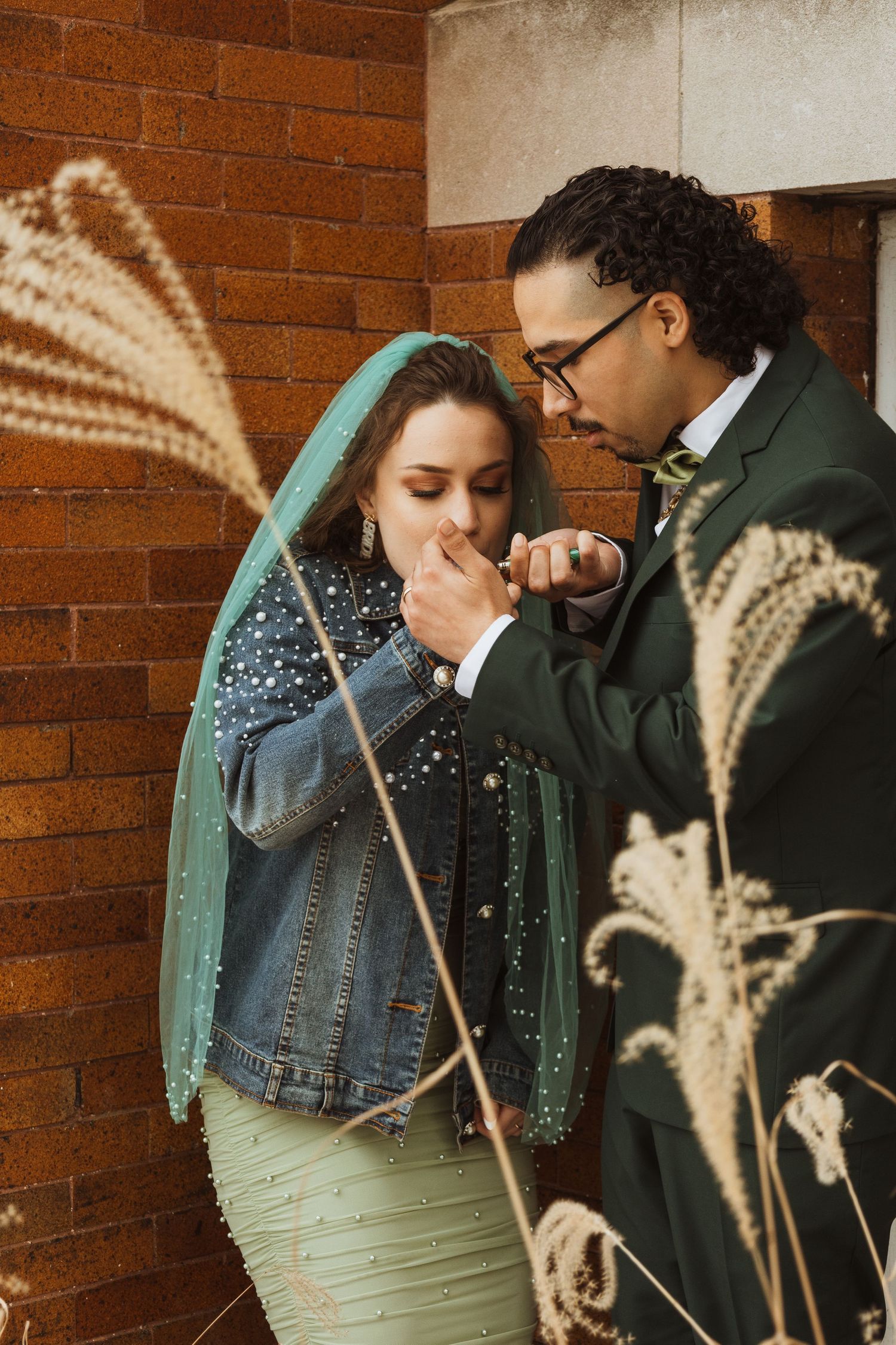 Groom in dark green suit kissing bride's hand outside brick wall, bride wearing teal veil.