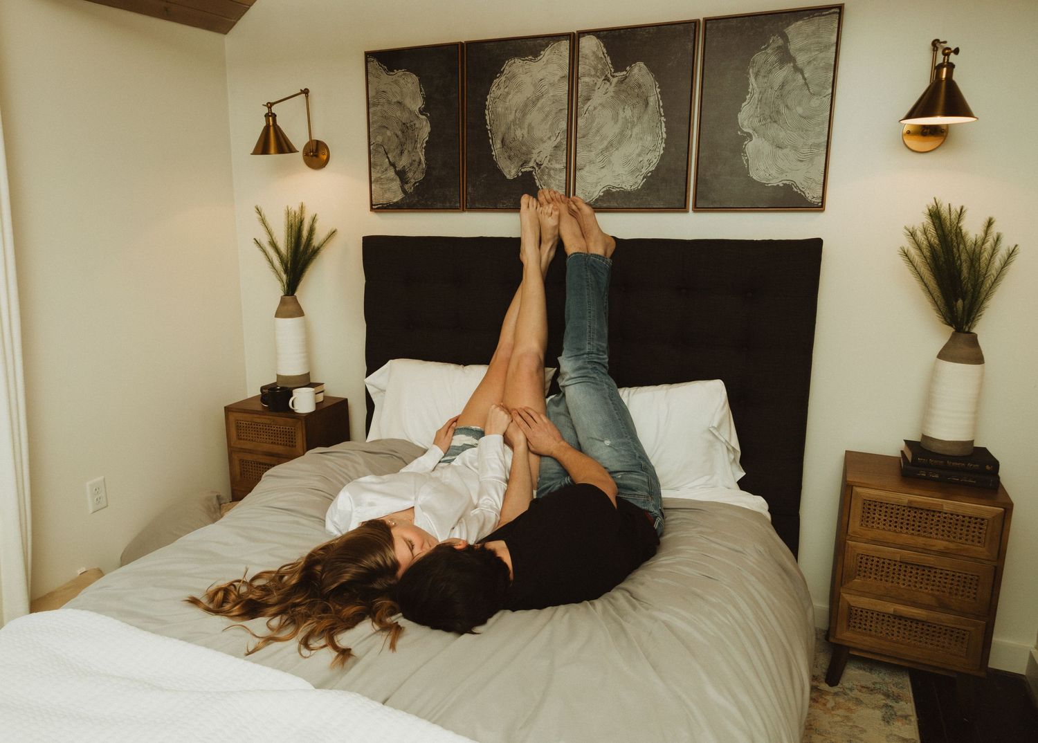 Two people playfully raising their legs while lying on a bed with cloud artwork above.