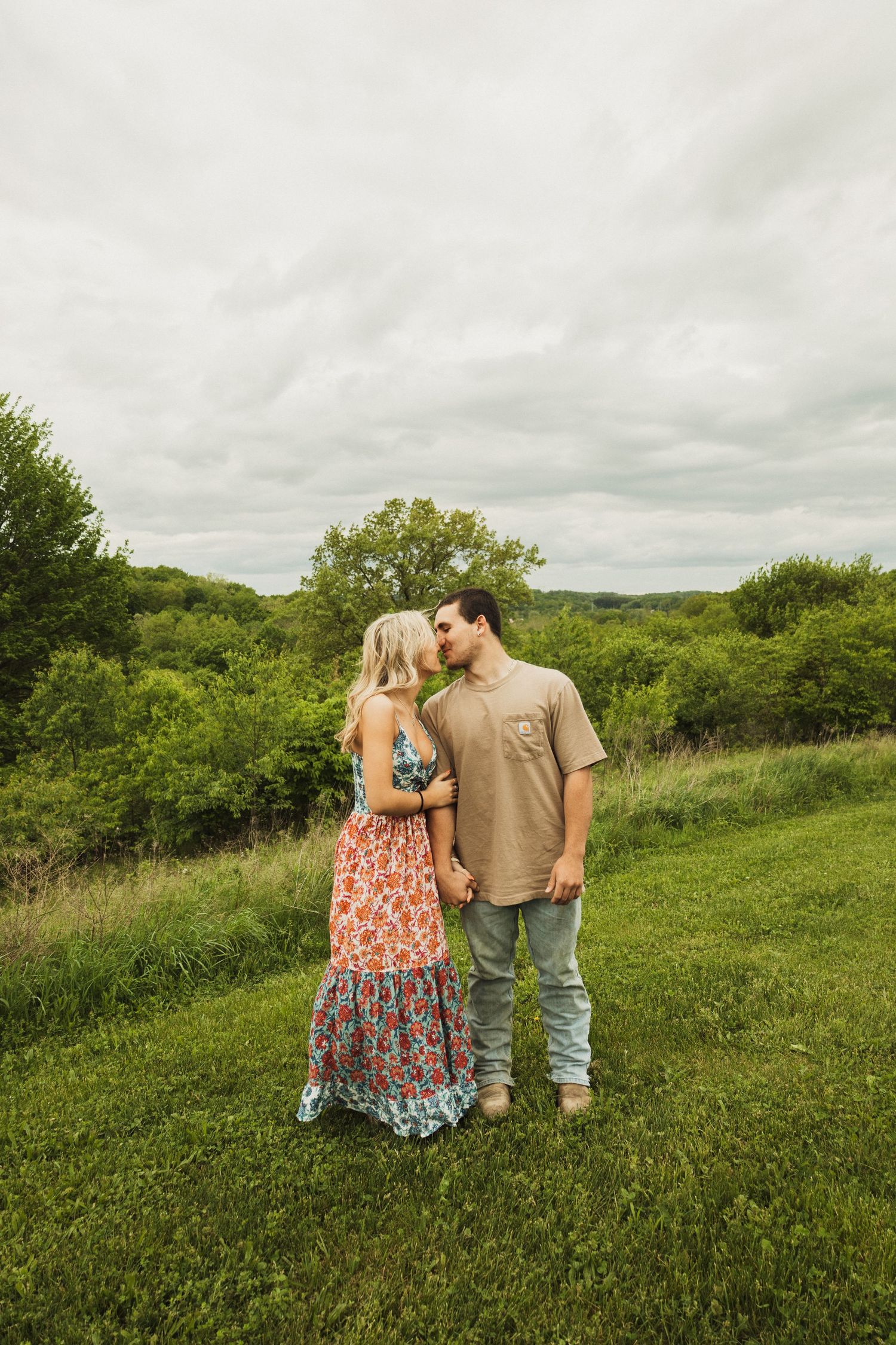 A couple stands together on a grassy hillside under cloudy skies, with her wearing a floral maxi dress.