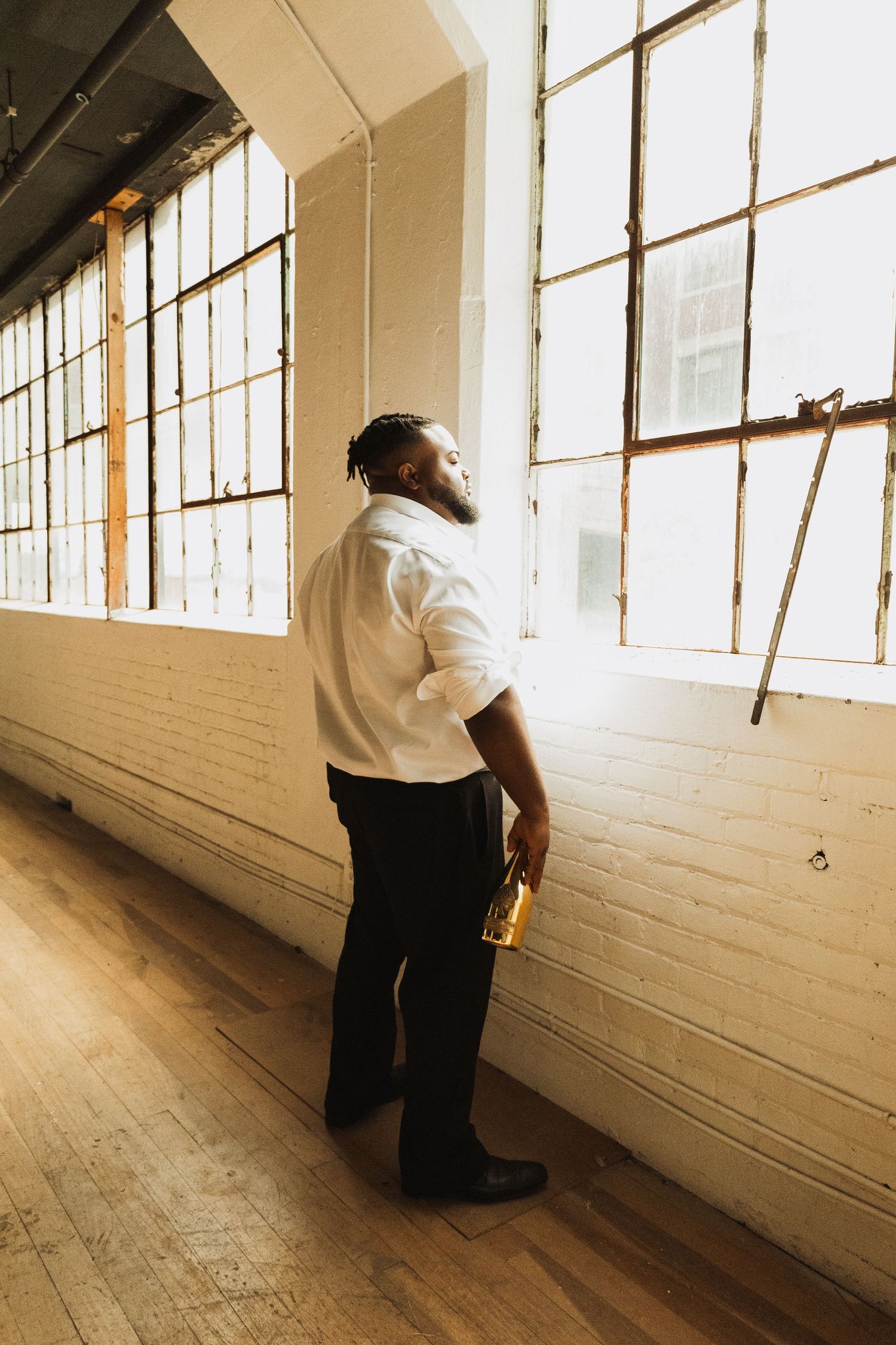 A person in a white shirt stands looking up at sunlight streaming through tall industrial windows in a hardwood-floored room.