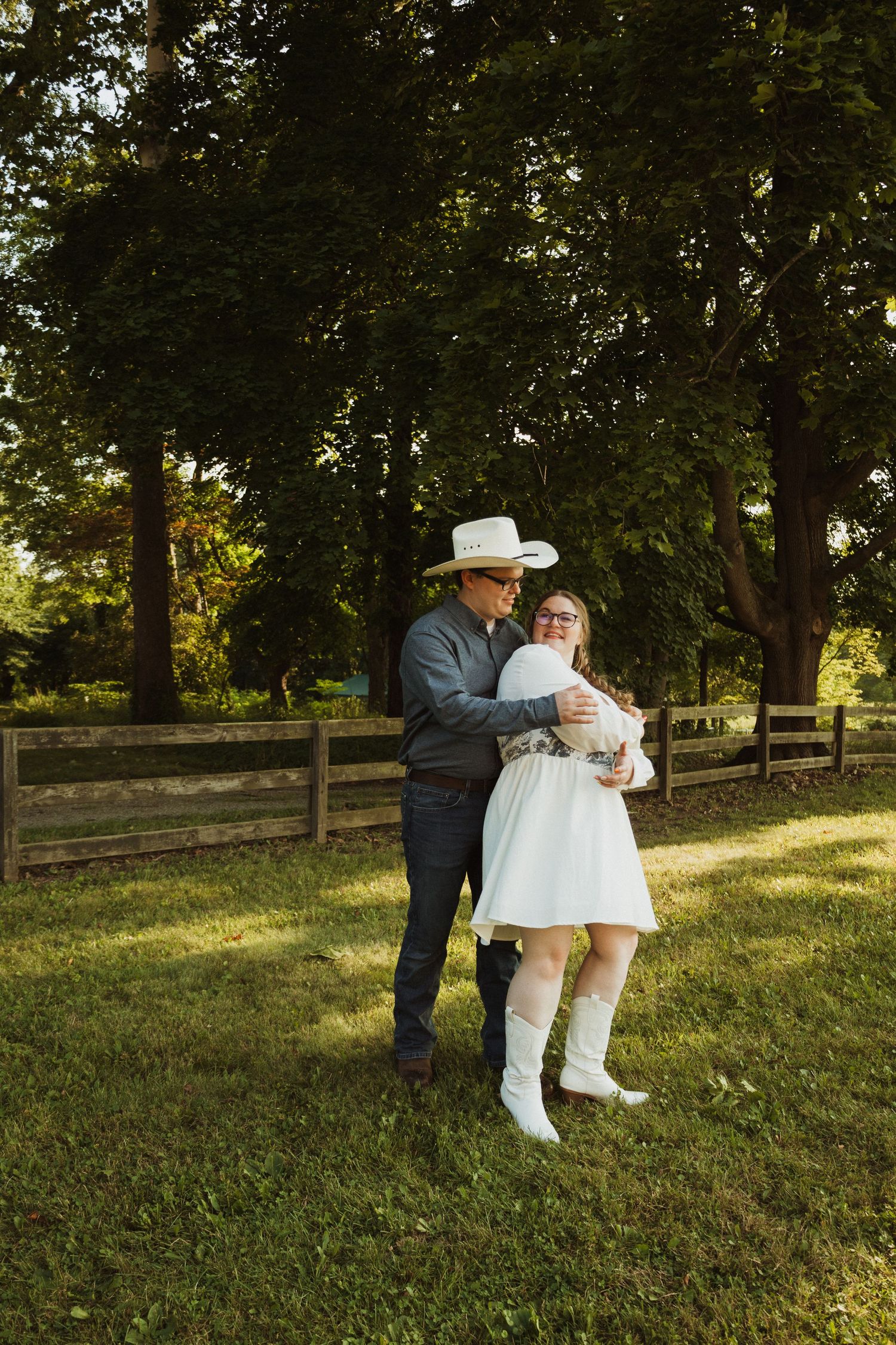 A couple embraces on a grassy slope with wooden fencing and trees in the background at sunset.