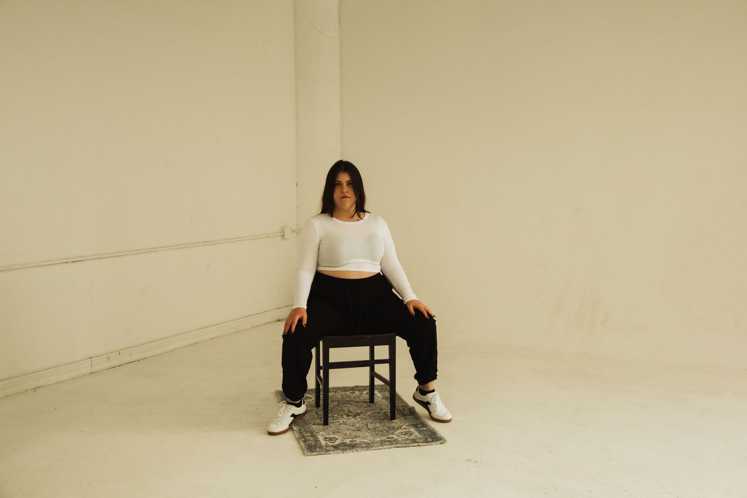 A person in dark pants and white shirt sits facing forward on a dark chair in an empty cream-colored studio space.