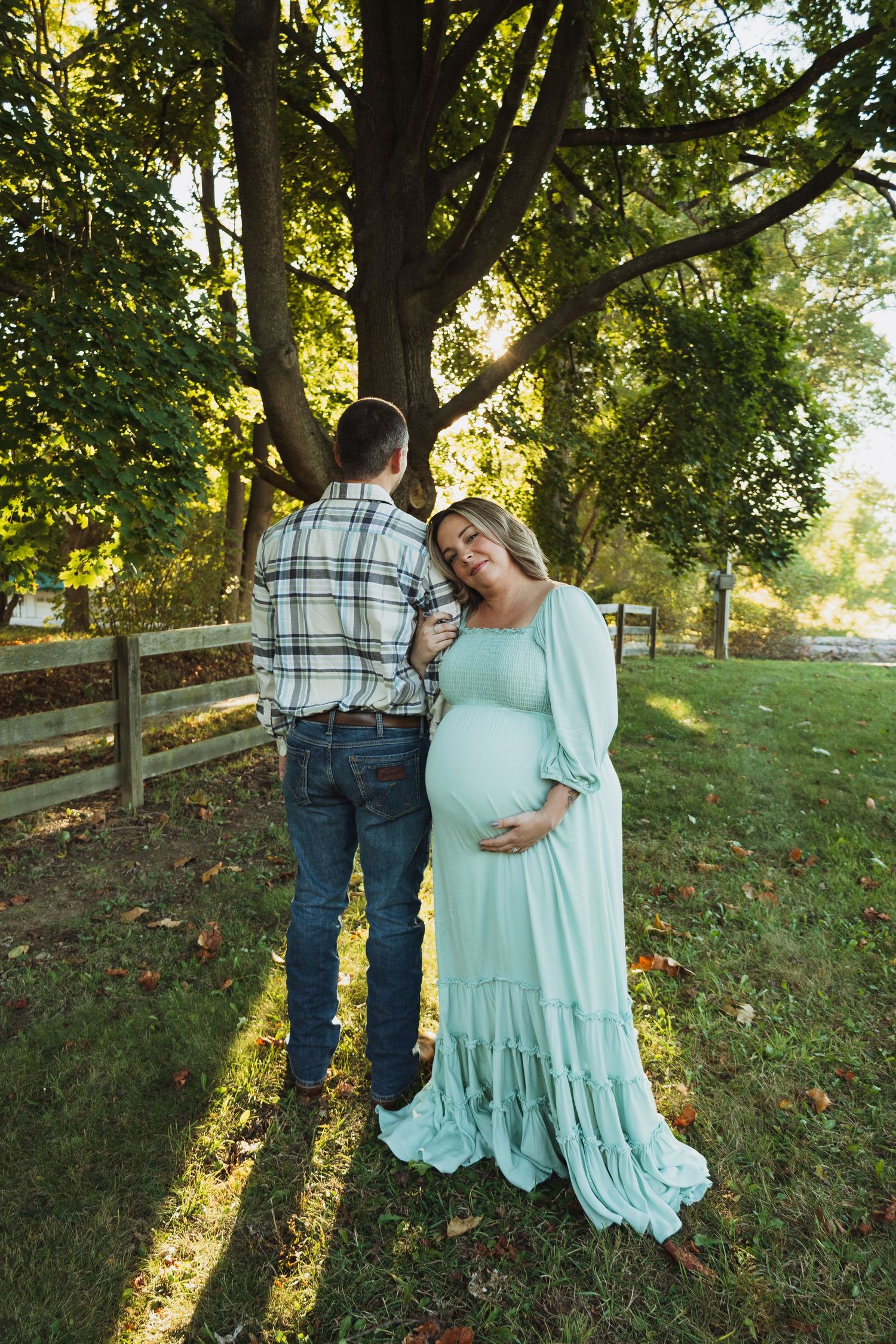 A couple stands under a sunlit tree in a park, with fall leaves scattered on the grass near a wooden fence.