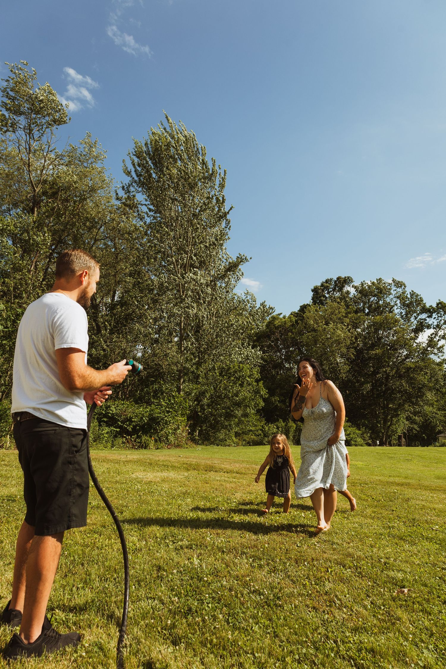 Family playing with garden hose on sunny day in green backyard, man spraying water while woman and child run toward him.