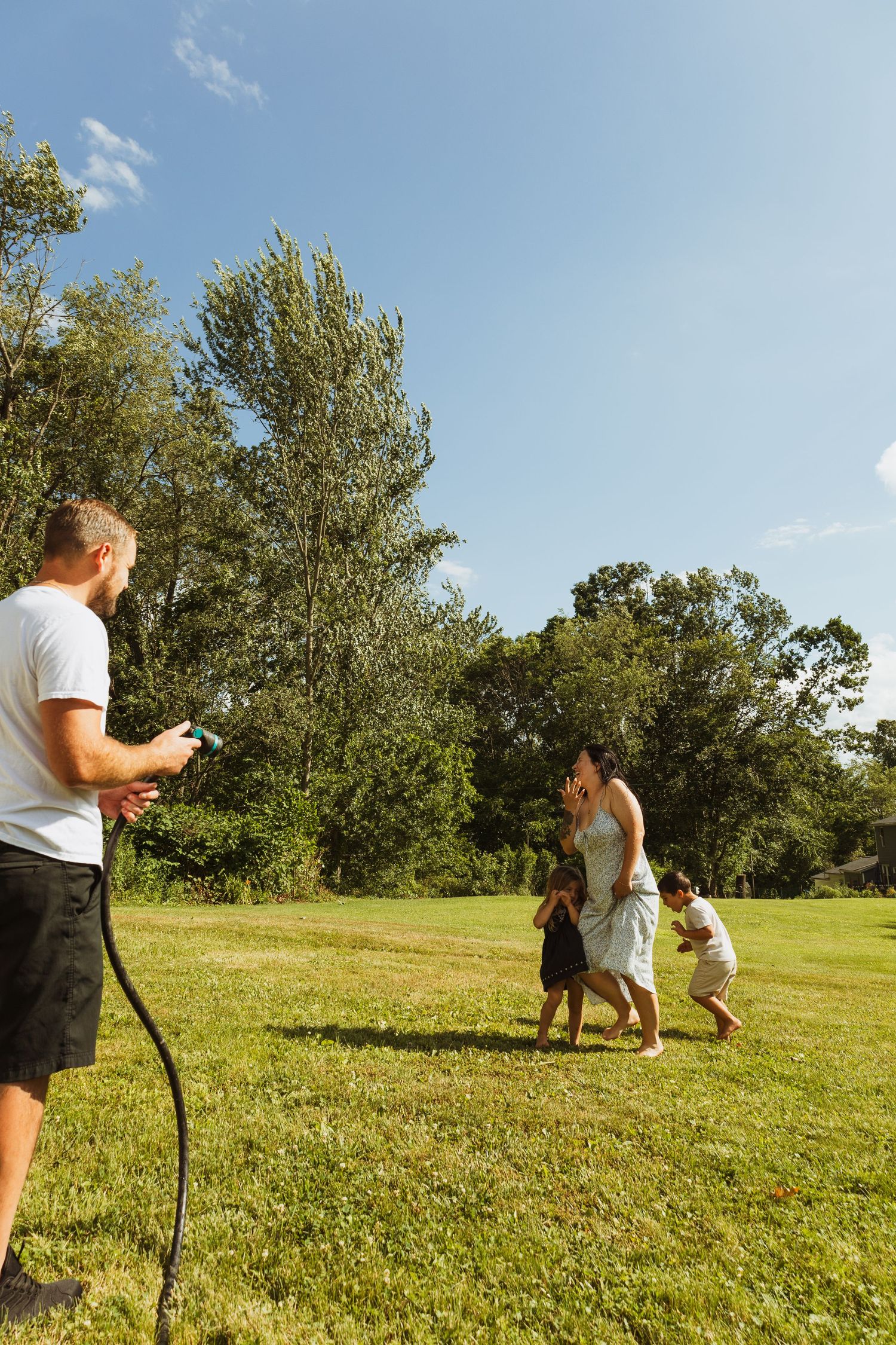 Family playing with a garden hose on a sunny day in a lush green backyard with trees and blue sky.