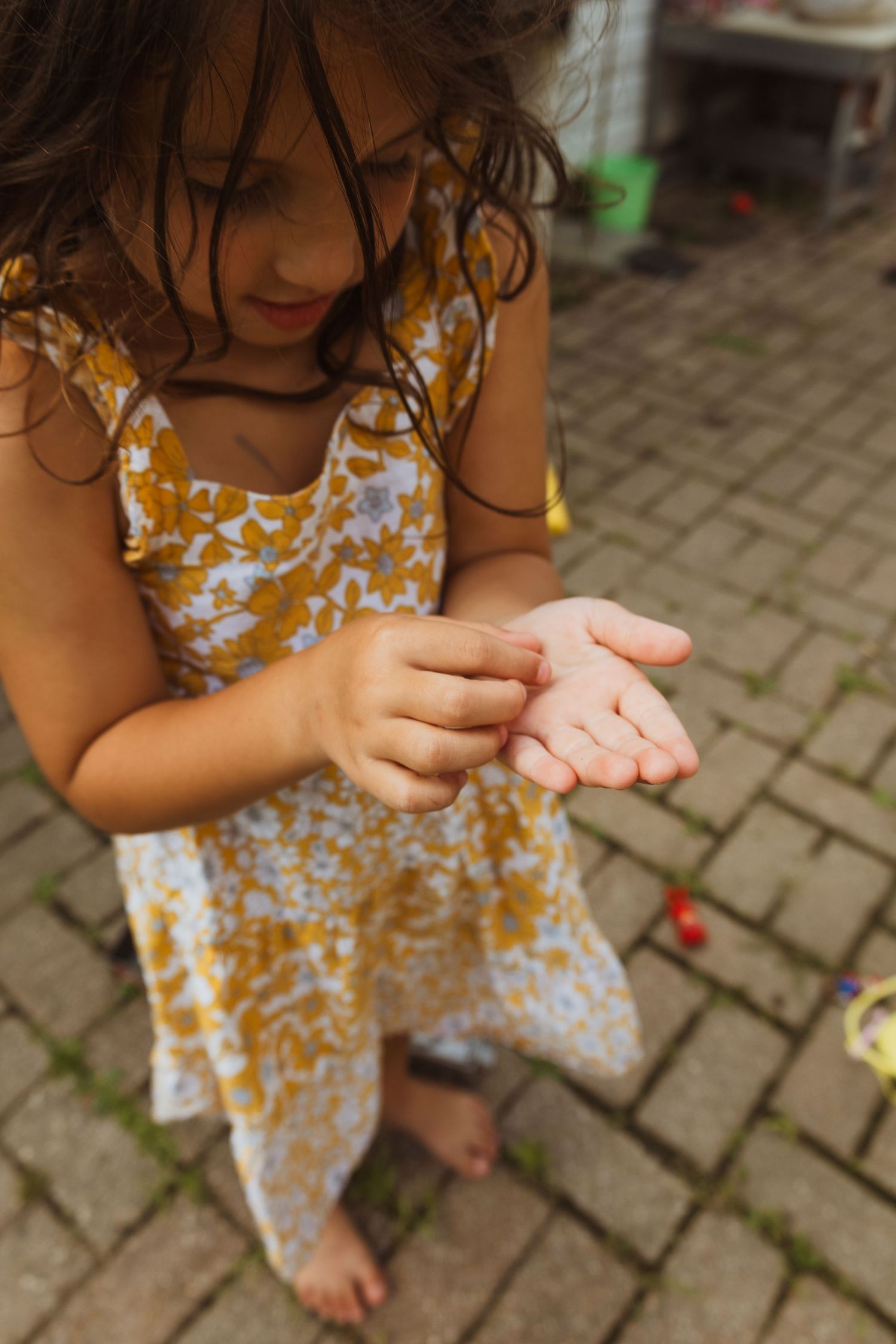 Young girl in floral dress examining her hands while kneeling on brick pavement outdoors.