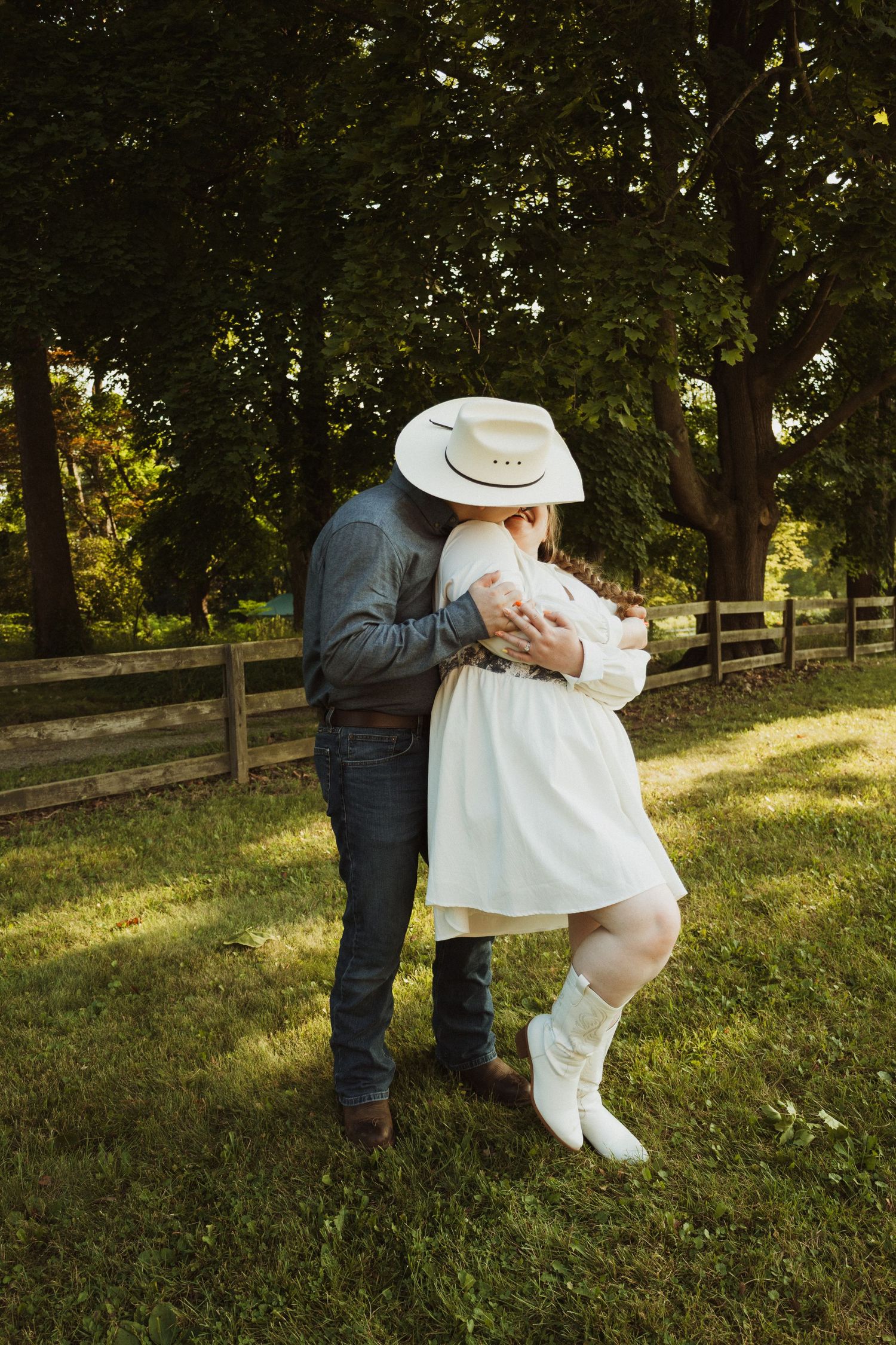 A romantic couple embraces on a sunny lawn with trees and wooden fencing in the background.