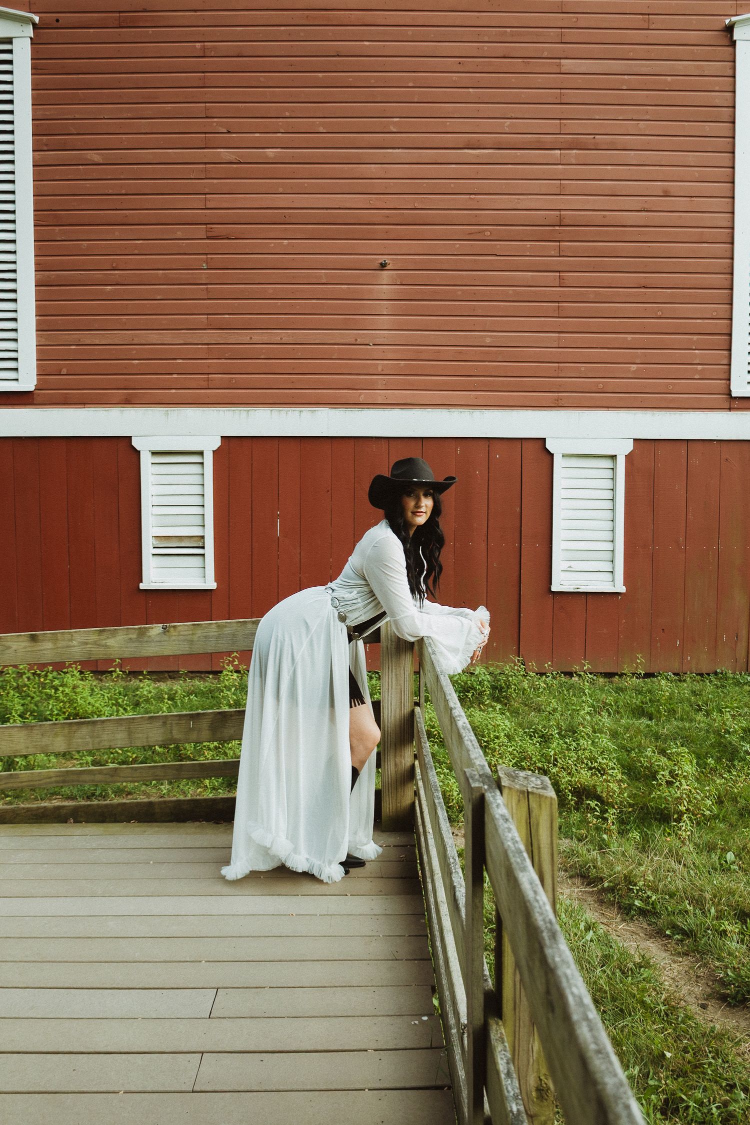 Figure in a long dress stands next to a rustic wooden fence and red barn wall.