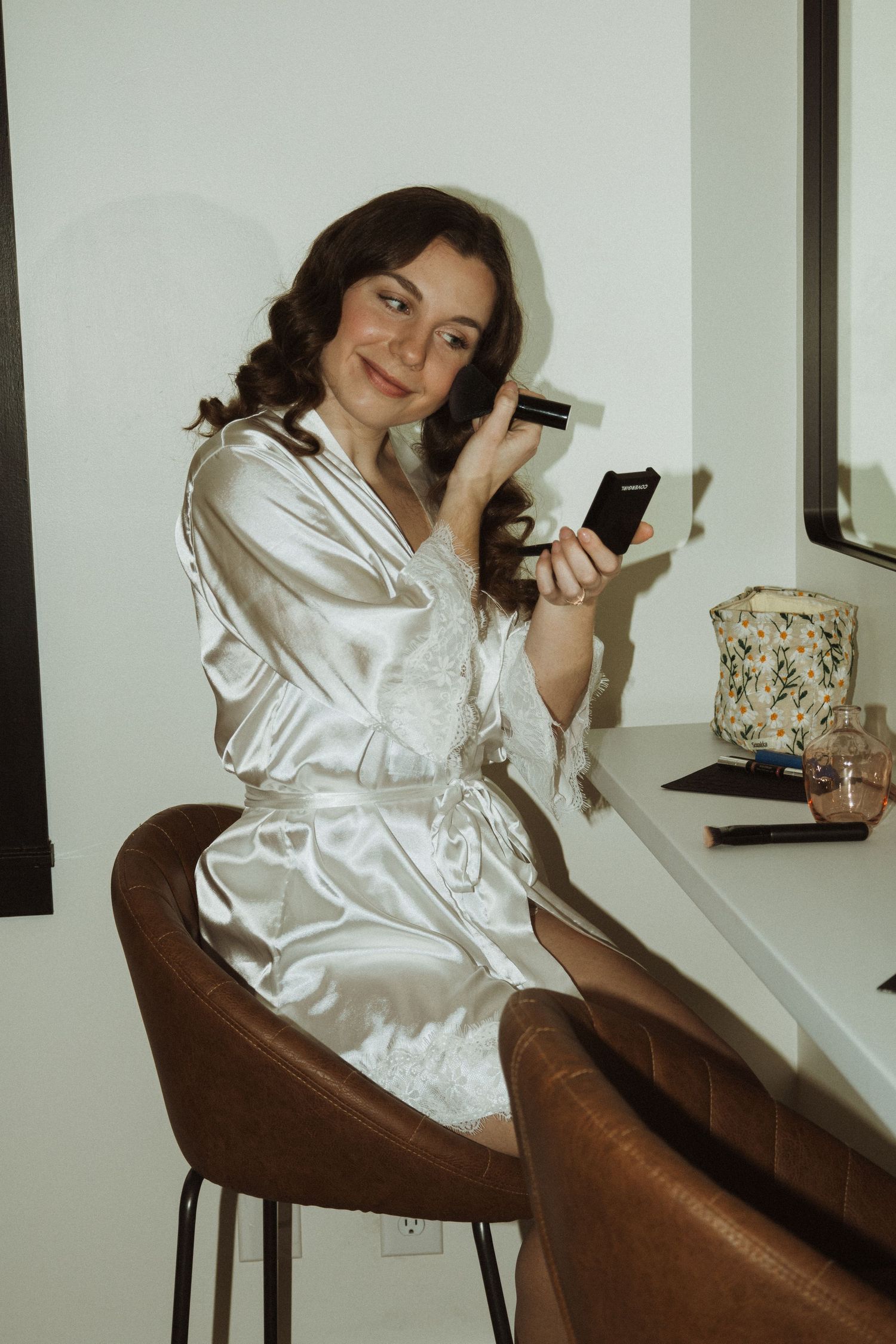 Person in white silk robe applies makeup while sitting at vanity mirror.