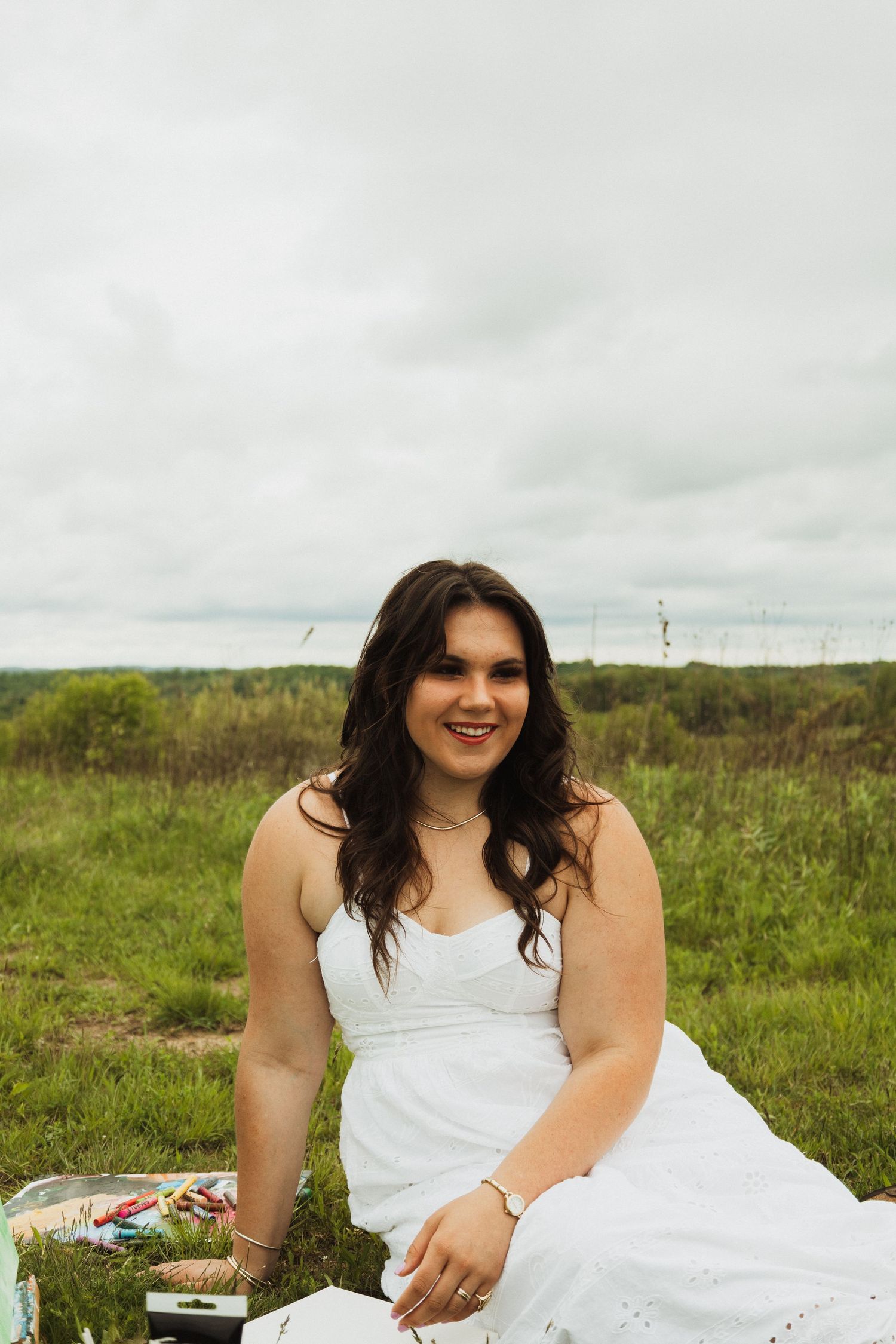 A person in a white dress sits on a blanket in a green field under an overcast sky.