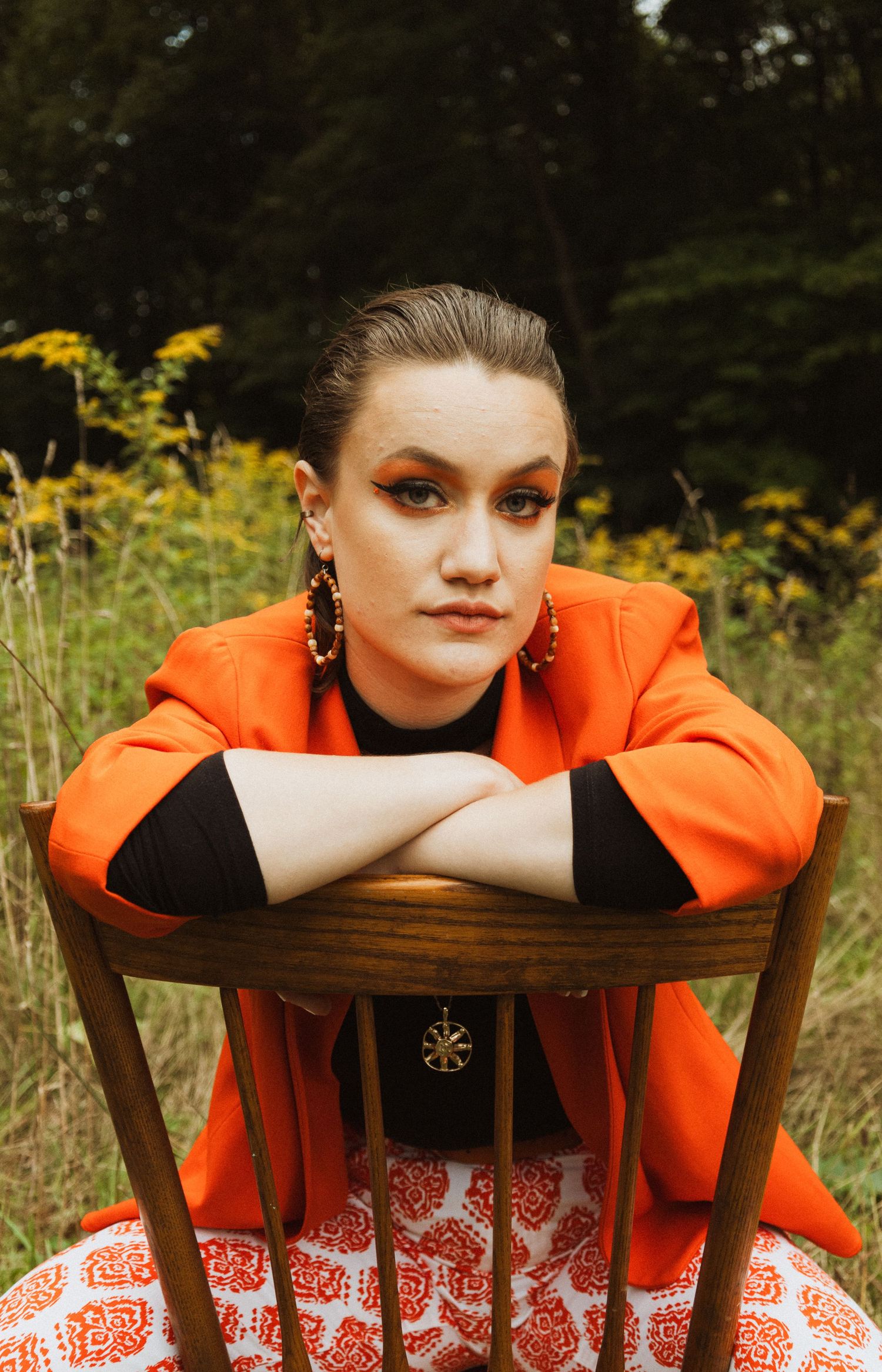 Close-up fashion portrait with dramatic makeup and orange blazer against wooden chair backdrop.