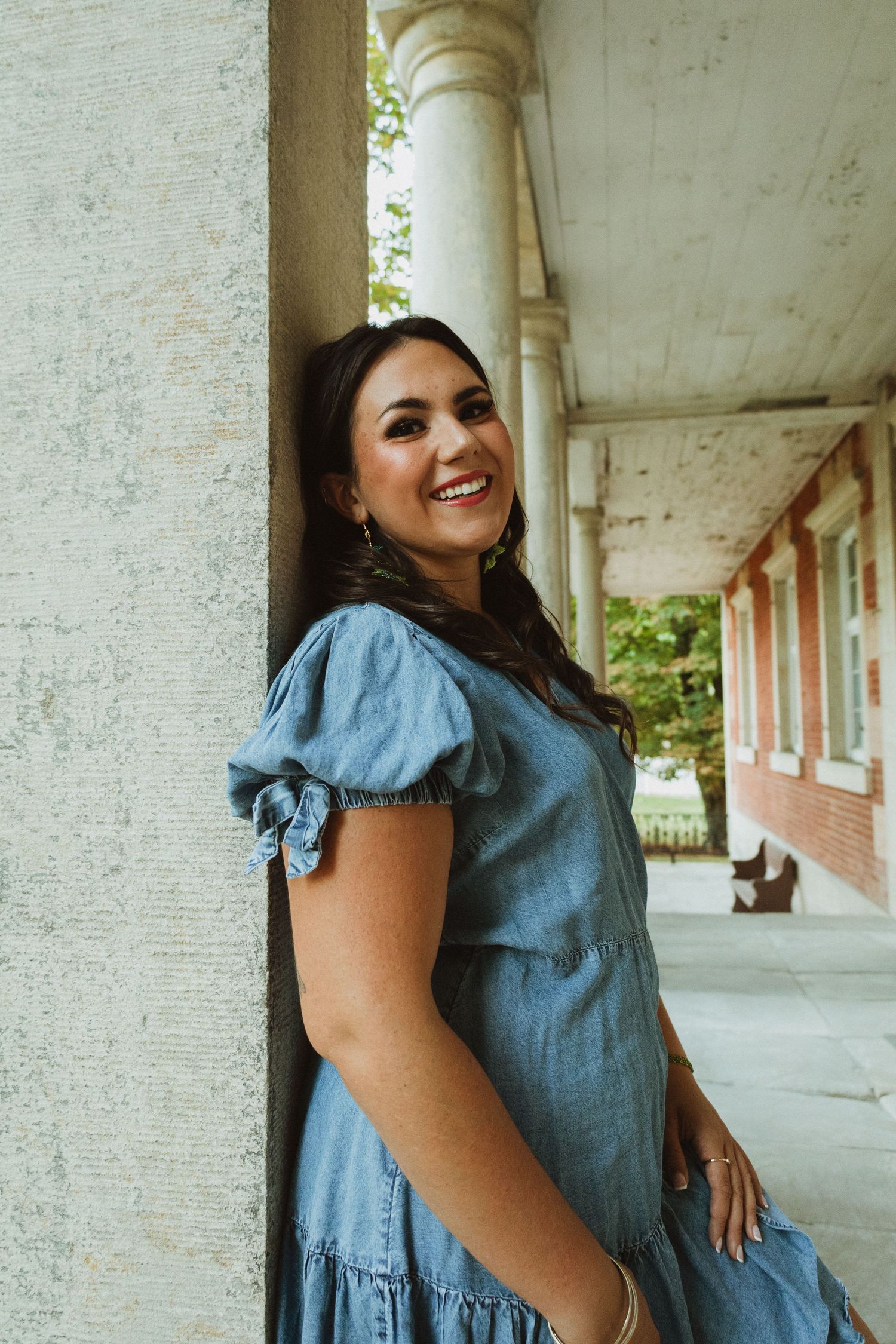 Person in a ruffled chambray dress poses playfully against white pillars on a covered porch.