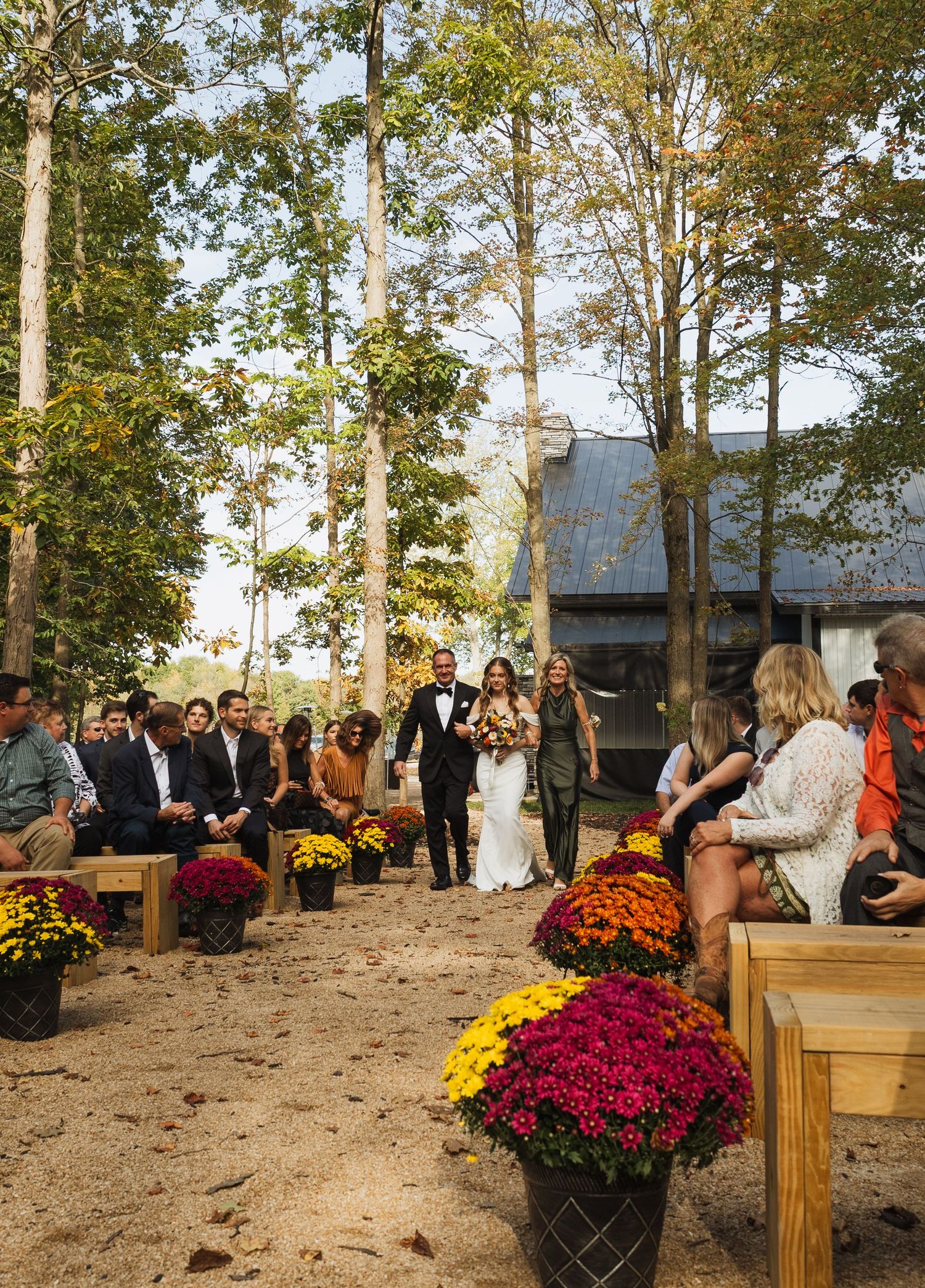 An outdoor wedding ceremony setup with wooden benches and colorful fall mums decorating a rustic woodland venue.
