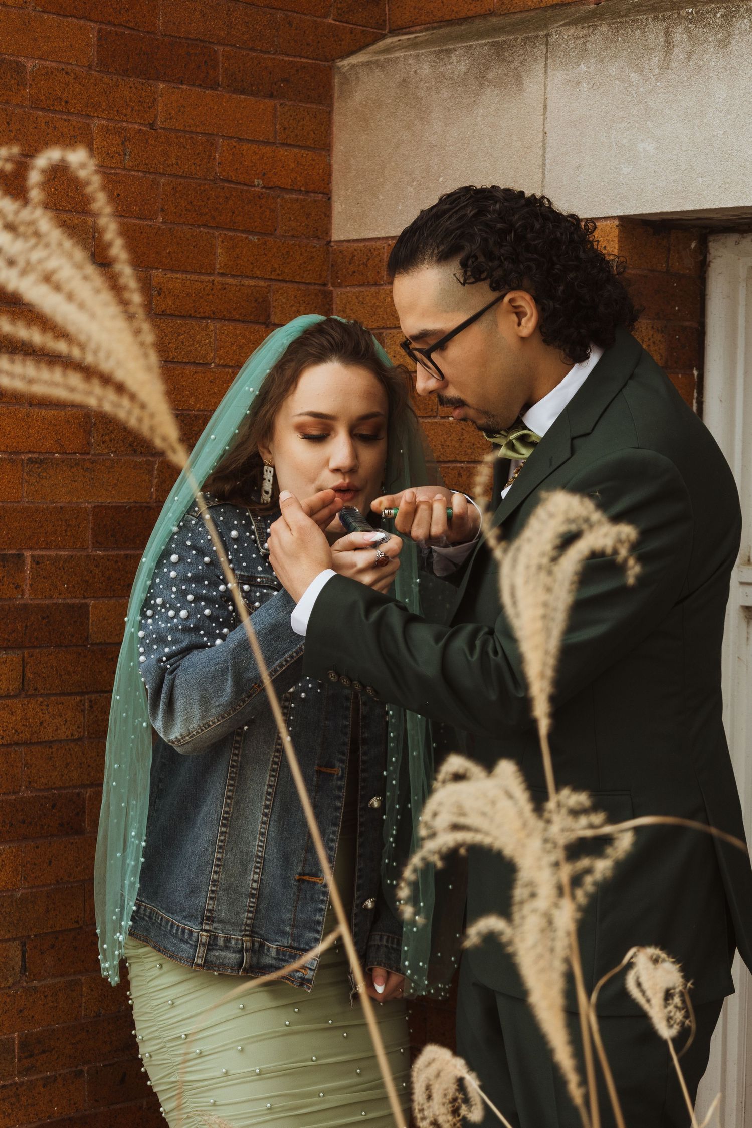 Bride and groom lighting cigarettes outdoors by brick wall, bride in green dress with teal veil and denim jacket.
