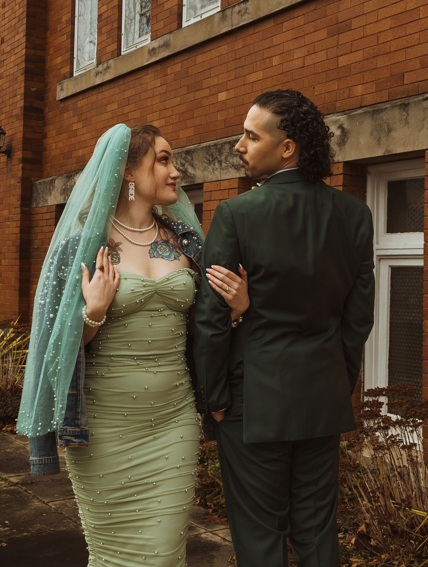 Bride in sage green pearl dress with teal veil and groom in dark green suit pose by brick building.