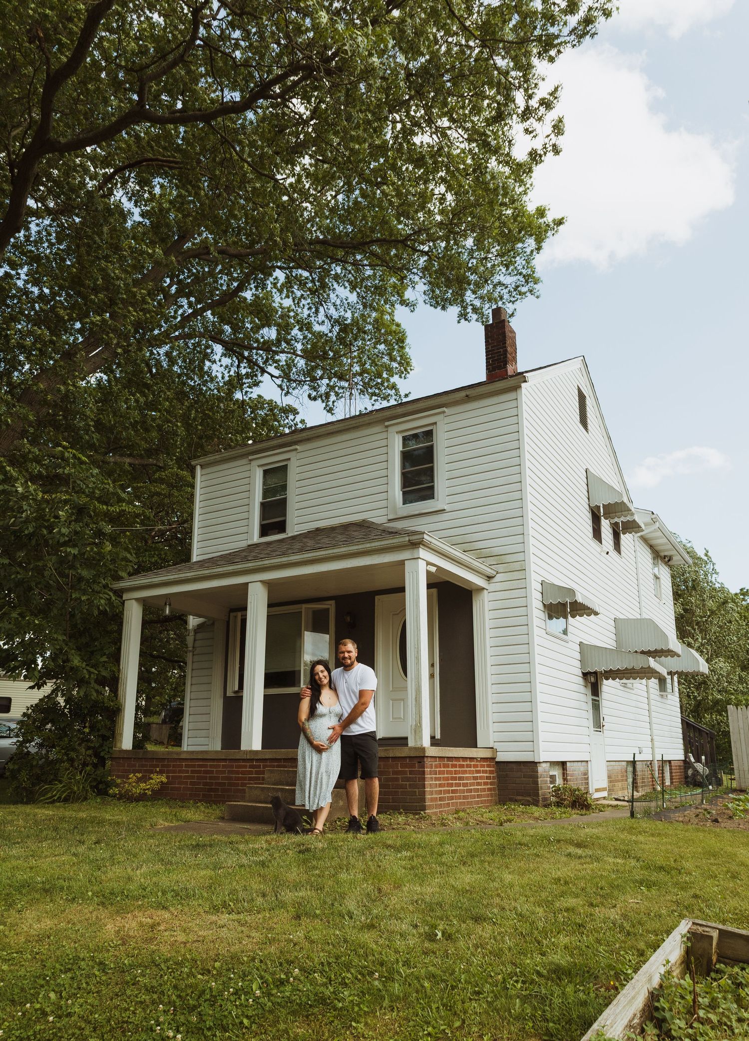 Couple standing in front of a white two-story colonial home with a covered porch surrounded by lush green trees.