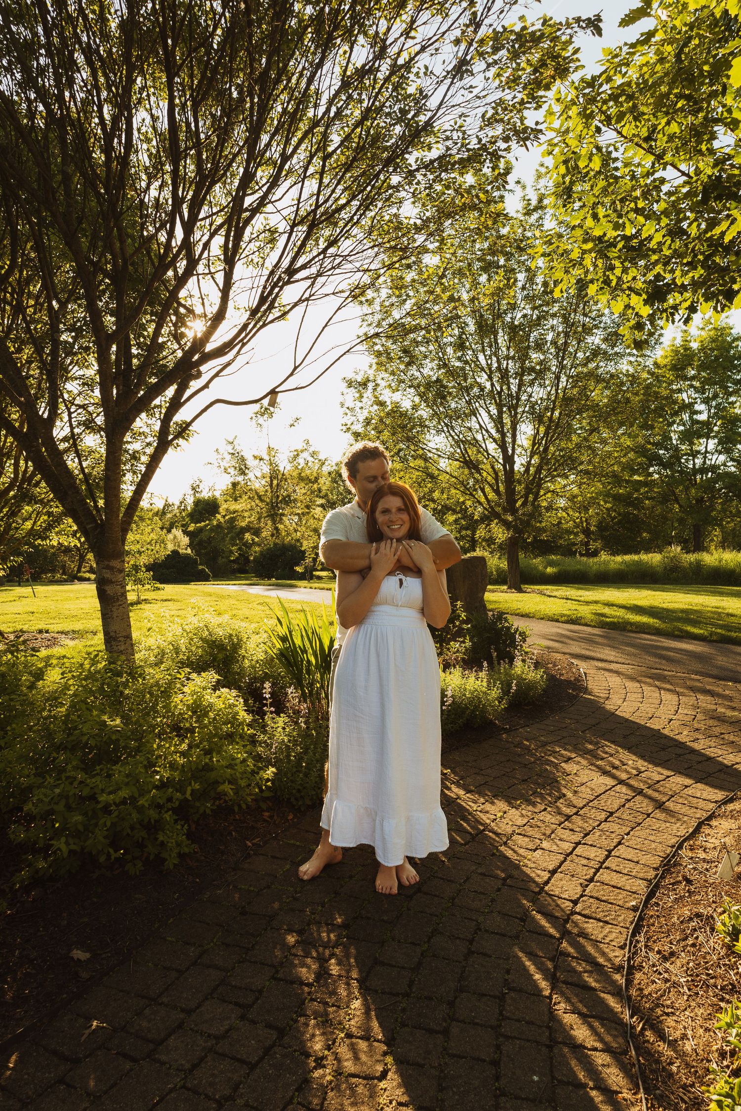 Couple embracing on a brick path in a sunlit garden, woman in white dress, surrounded by lush green trees.