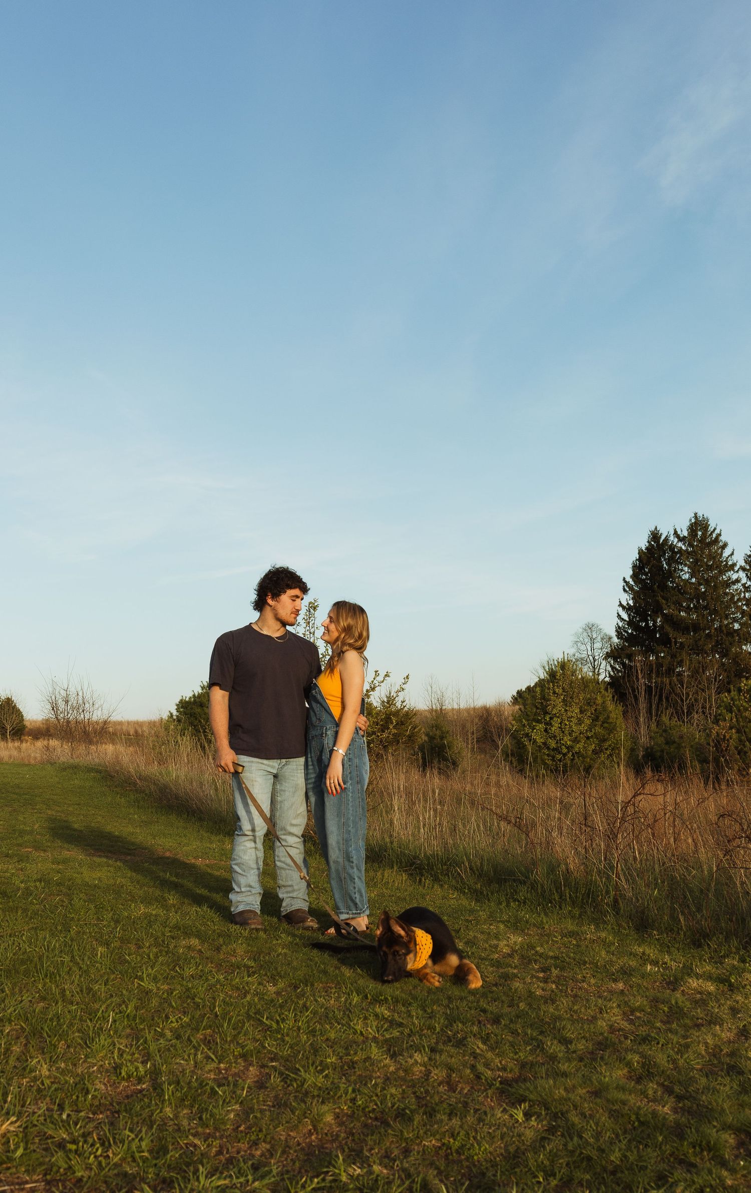 Couple standing together in a sunny field with their dog, surrounded by green grass and trees under a clear blue sky.