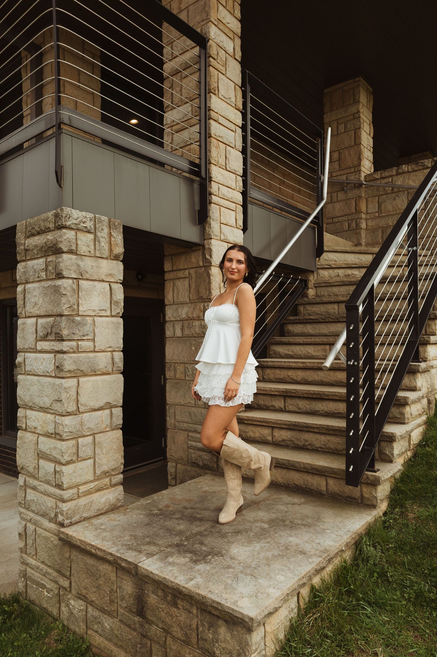 Woman in white dress and cowgirl boots posing on stone steps of a modern building with cable railings.
