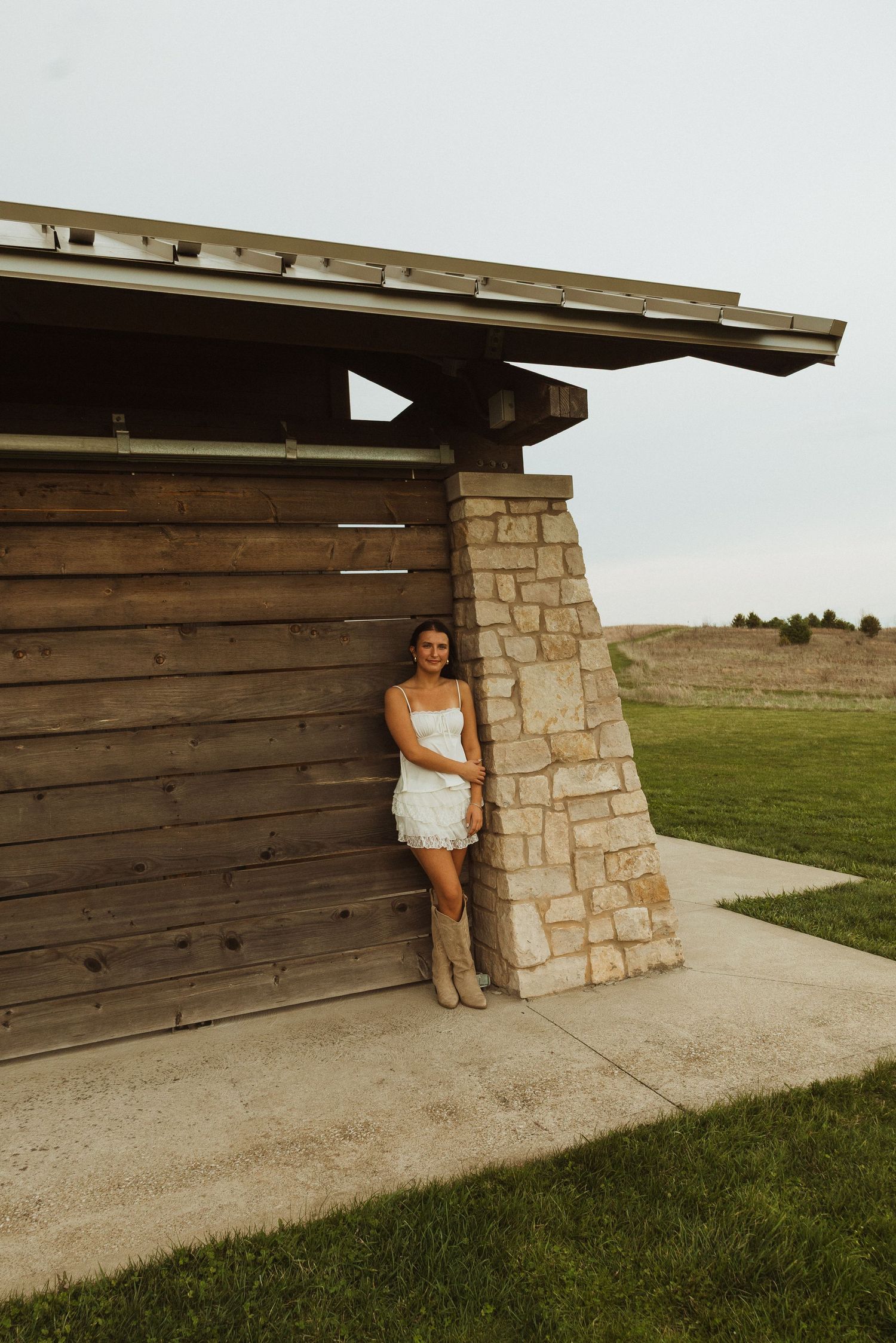 Woman in white dress and cowboy boots leaning against rustic wood and stone building on a green lawn.