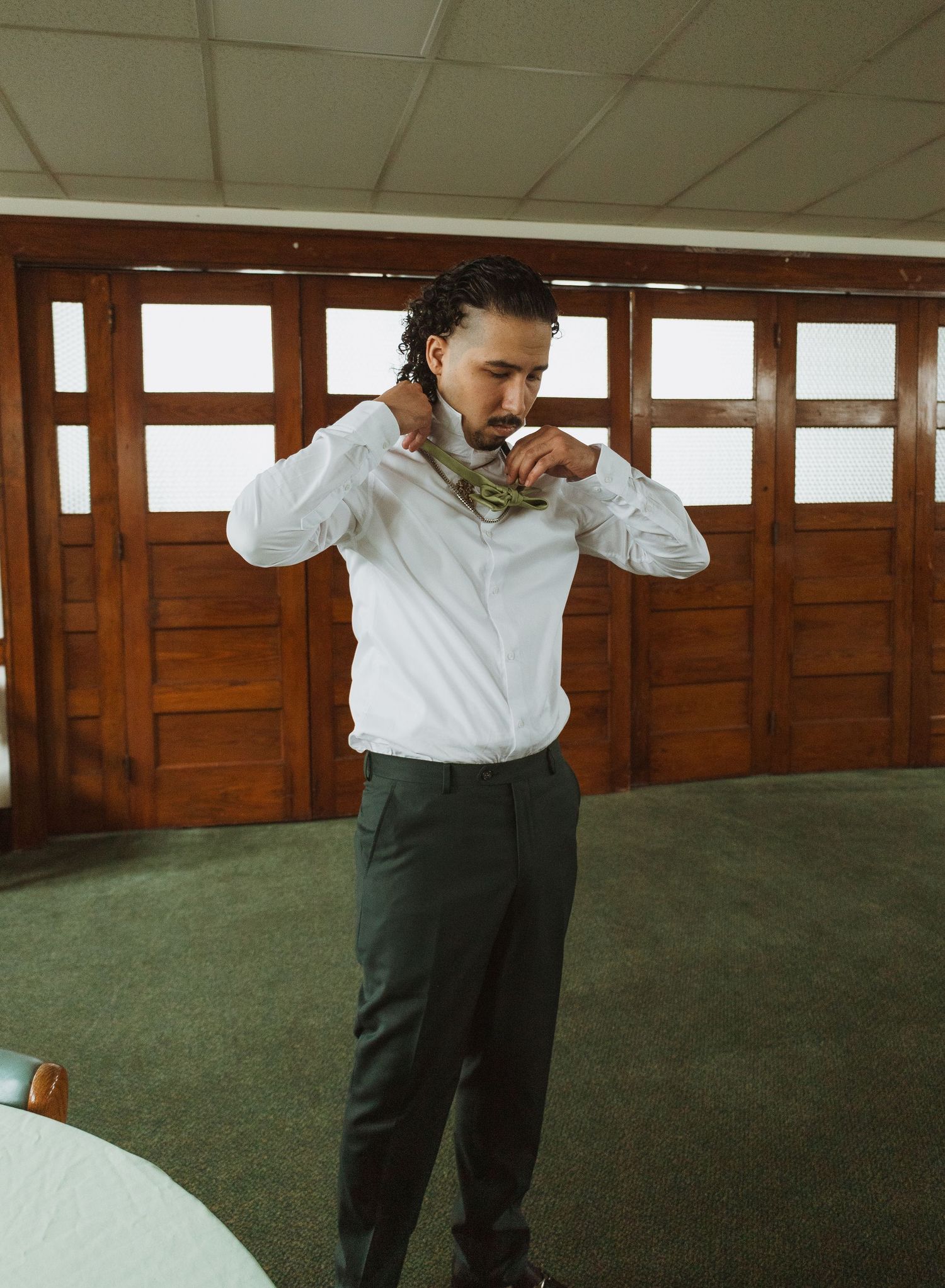 Groom in white shirt and gray pants adjusting his green floral bow tie before the wedding ceremony.