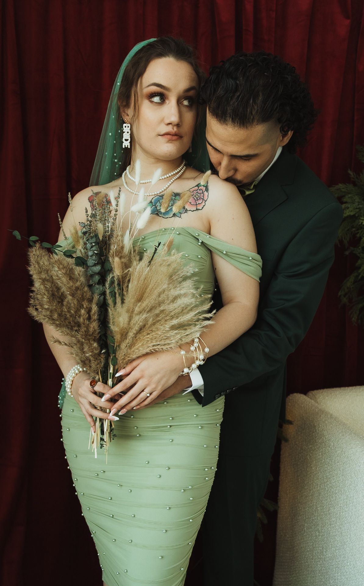 Bride in mint green pearl gown with veil and tattoos holds dried bouquet as groom in dark suit kisses her shoulder.