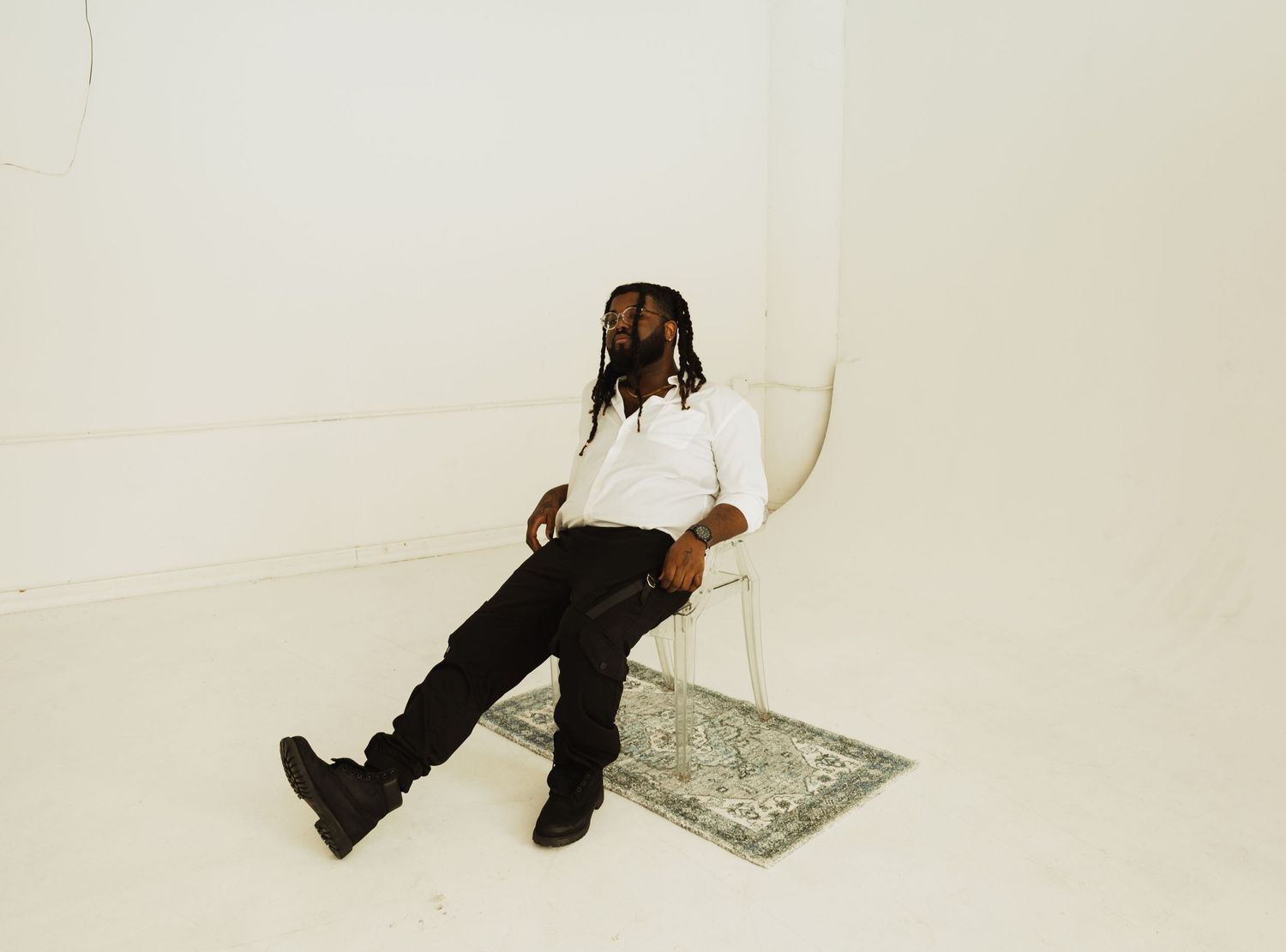 Person with braids wearing a white shirt and black pants sits casually in a white chair against a plain background.