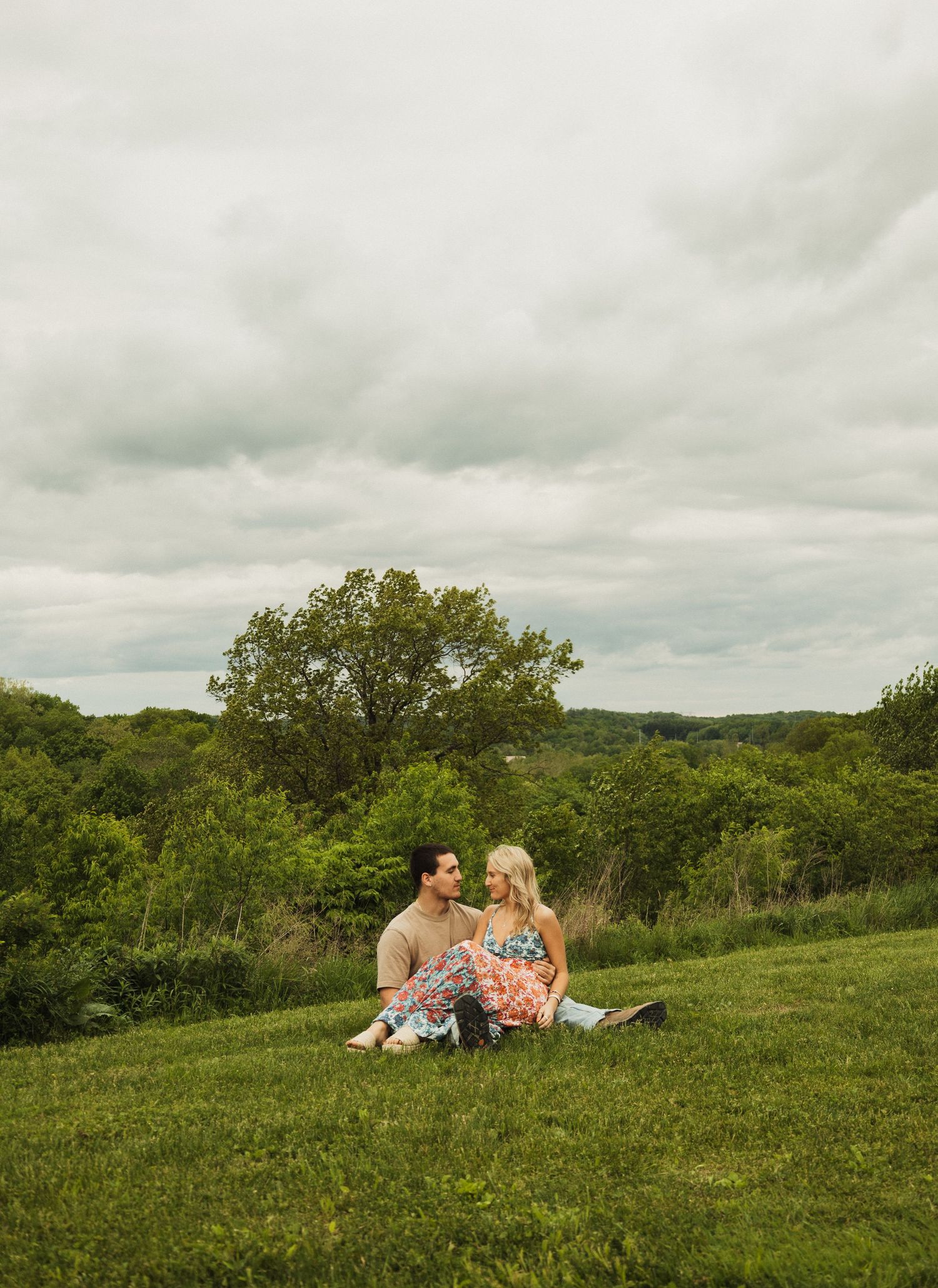A couple sits together on a grassy hillside overlooking a scenic wooded landscape.