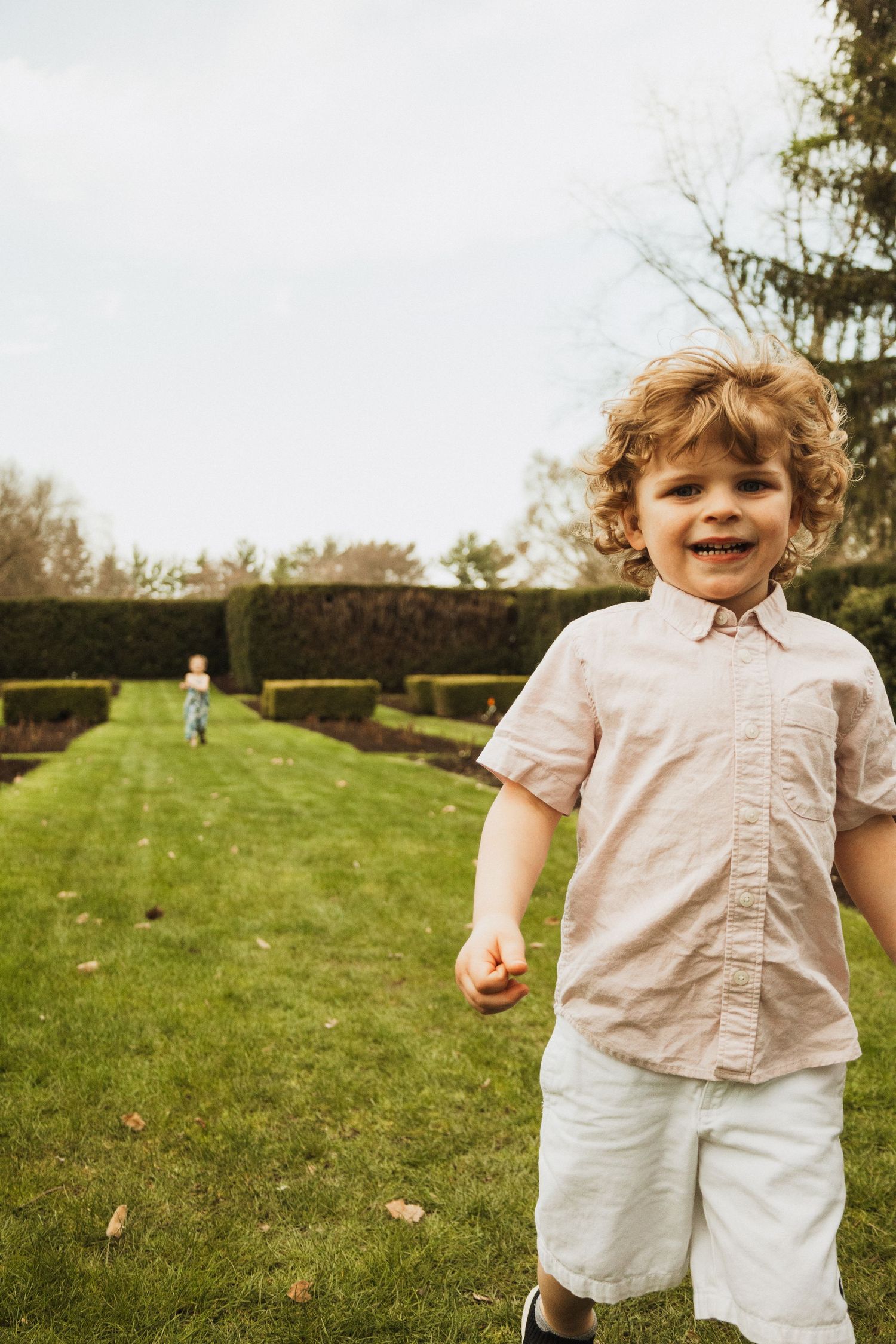 Young child in light colored clothing smiling at camera on a green lawn with trees in background.